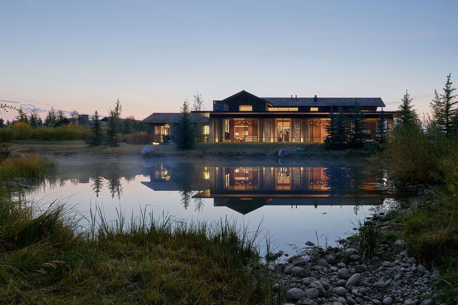 Twilight view of a modern waterfront home with illuminated glass windows perfectly reflecting in a calm pond at dusk.