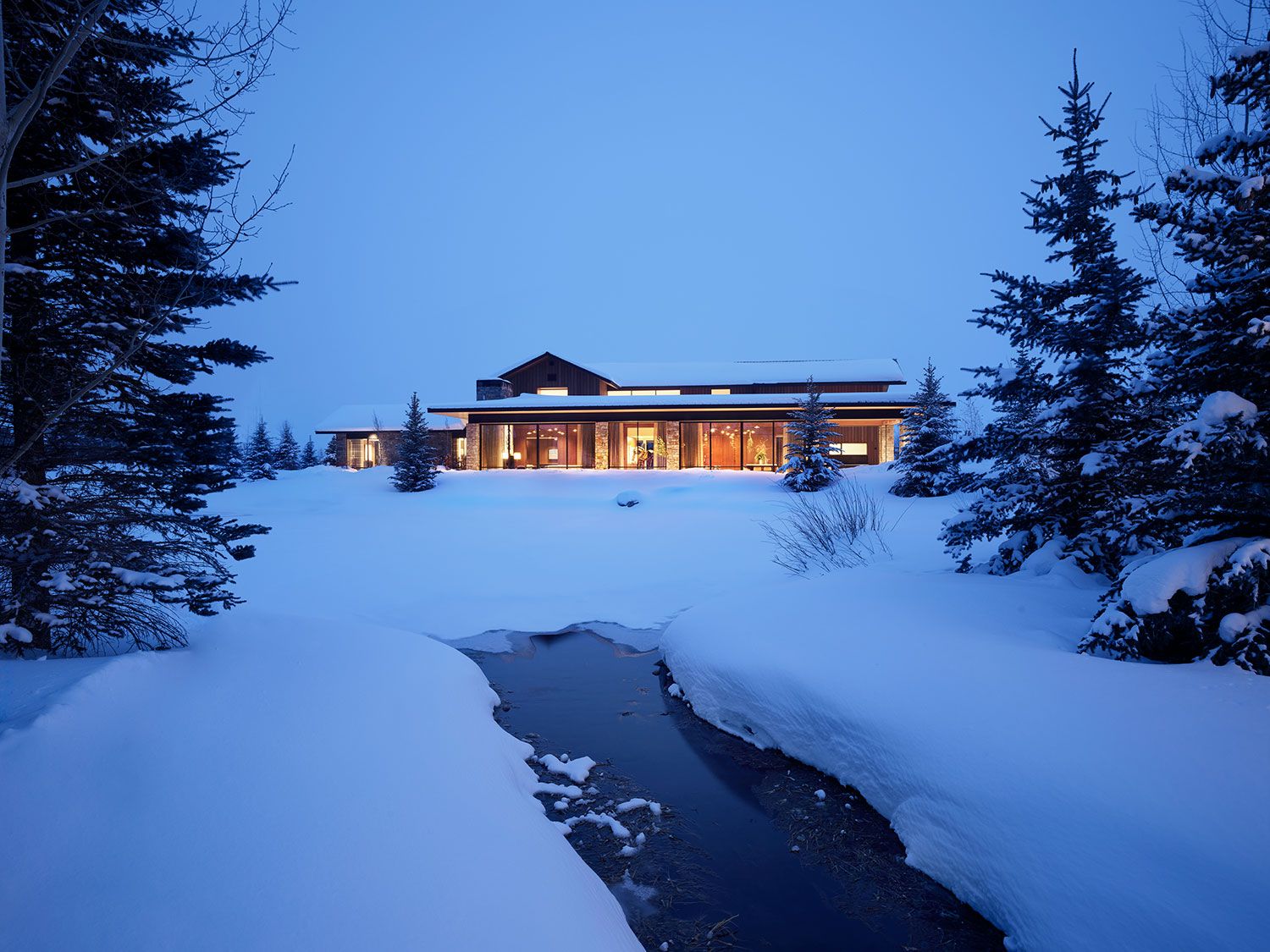 Twilight view of a luxury modern mountain home illuminated from within, nestled in a snowy landscape with a small unfrozen stream in the foreground.