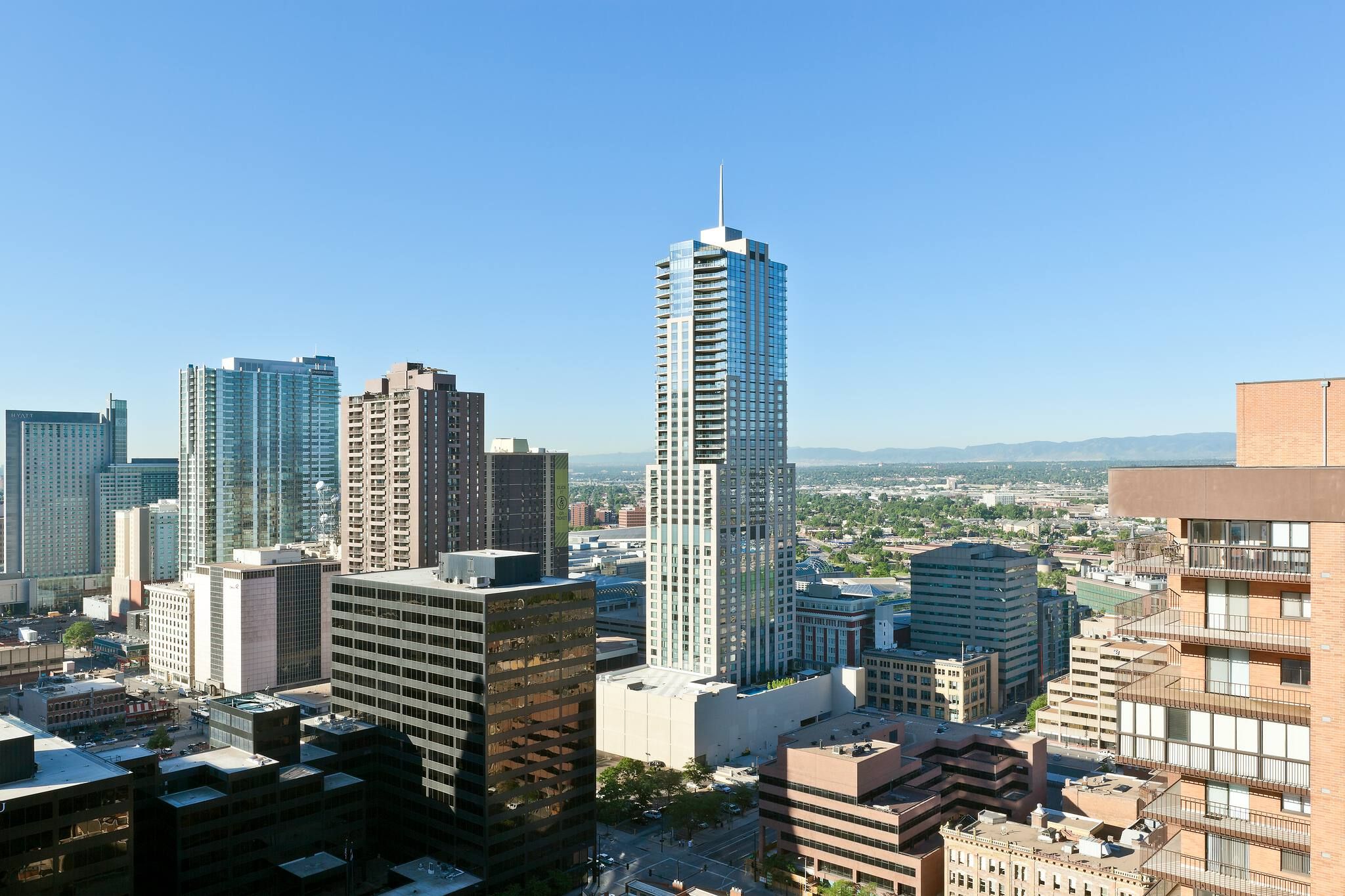 Downtown city skyline dominated by a sleek, modern glass skyscraper with a spire, surrounded by a mix of office and residential buildings, with distant mountains visible under a clear blue sky.