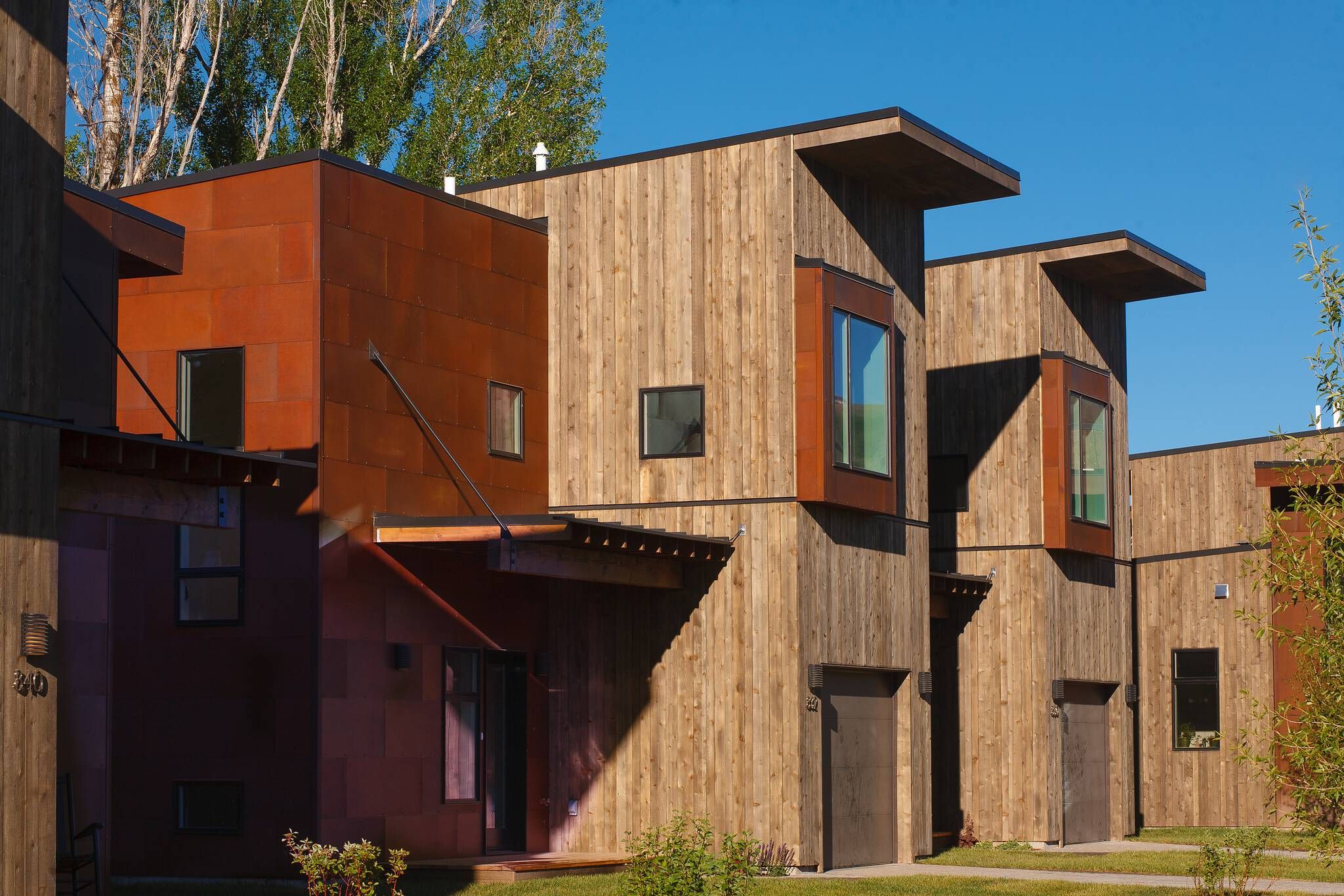 Modern townhouses featuring natural wood siding and rusted metal panels, with flat roofs, deep overhangs, and large geometric windows. Bright sunlight creates strong shadows on the facades under a clear blue sky.