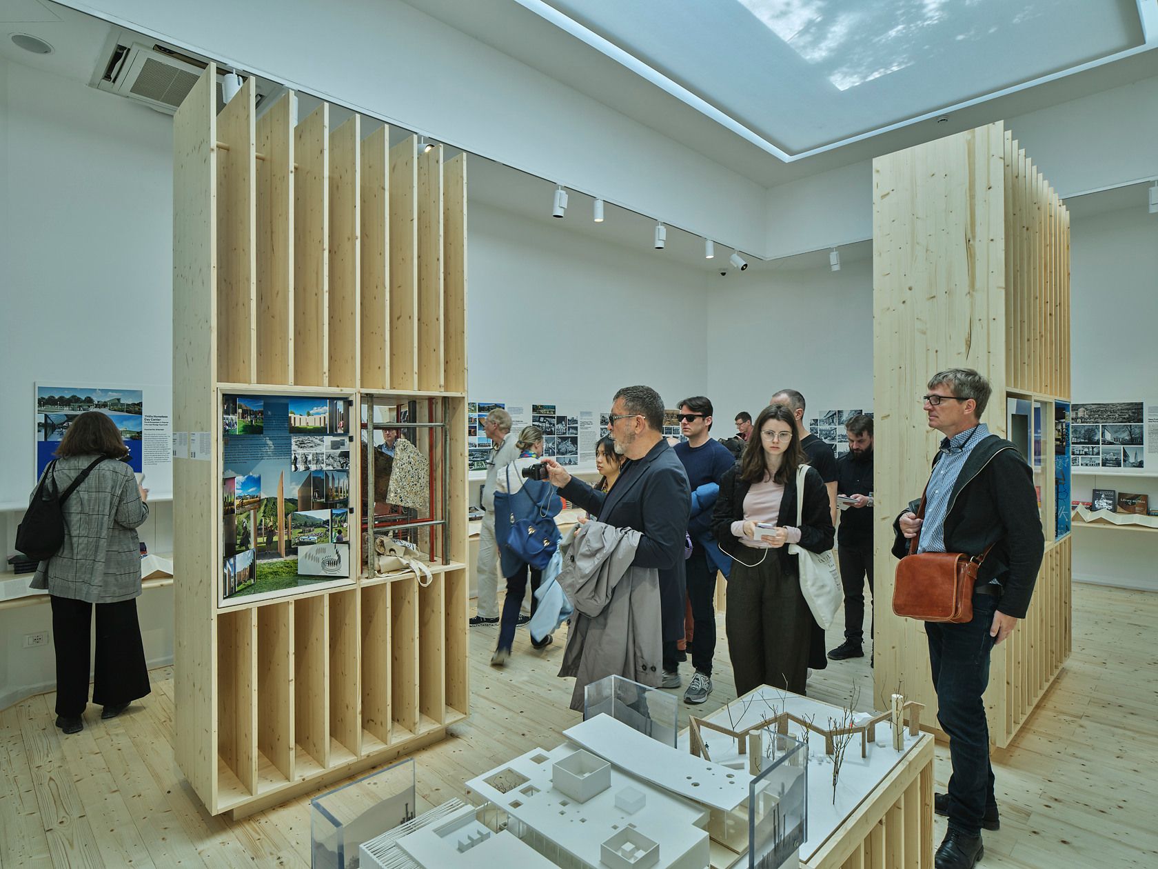 Visitors explore a modern architectural exhibition featuring bright wooden display units, scale models, and wall posters in a well-lit gallery space.