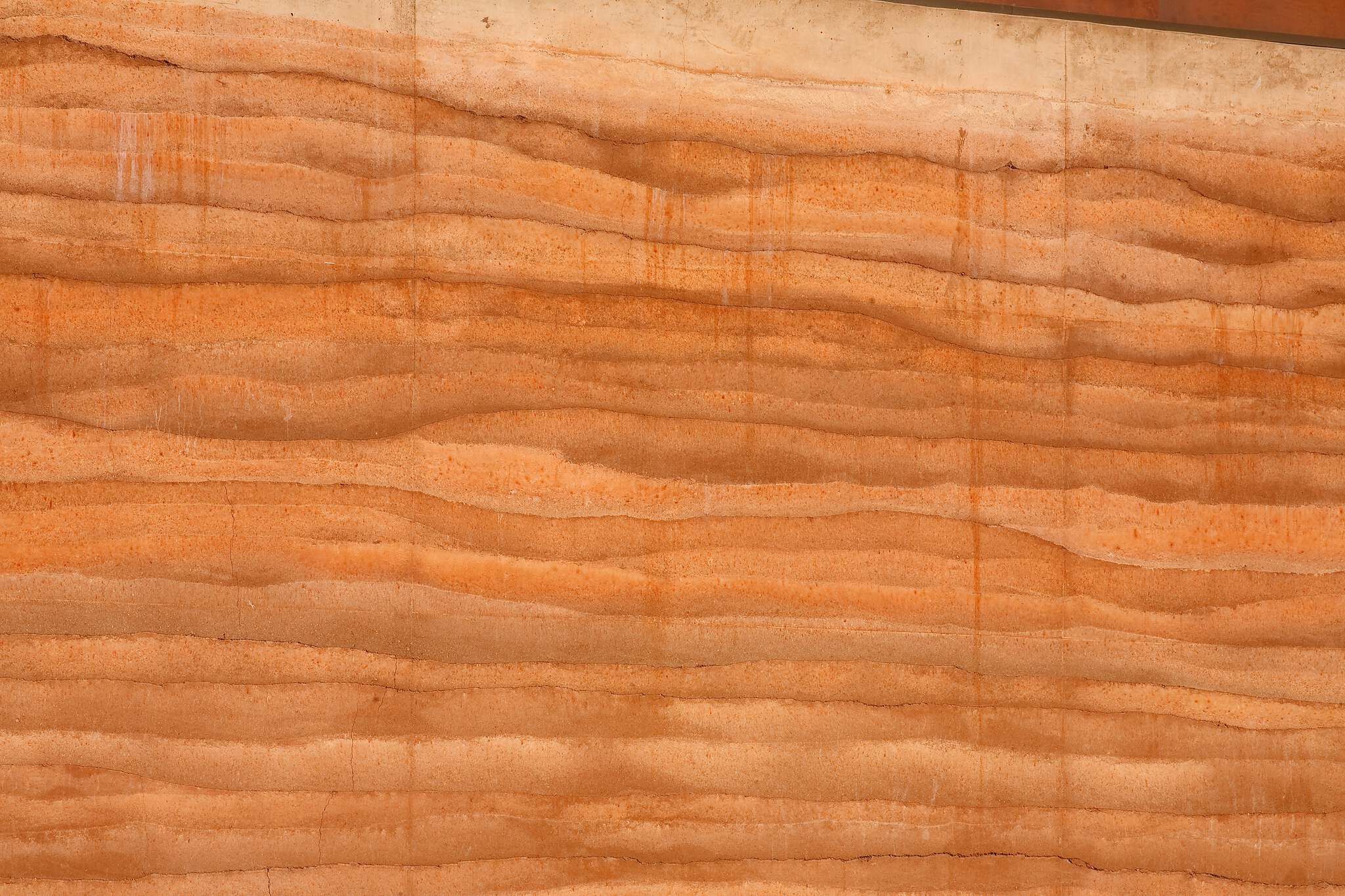 Textured rammed earth wall with distinct, undulating horizontal layers in warm reddish-brown and terracotta tones, topped by a light concrete section.