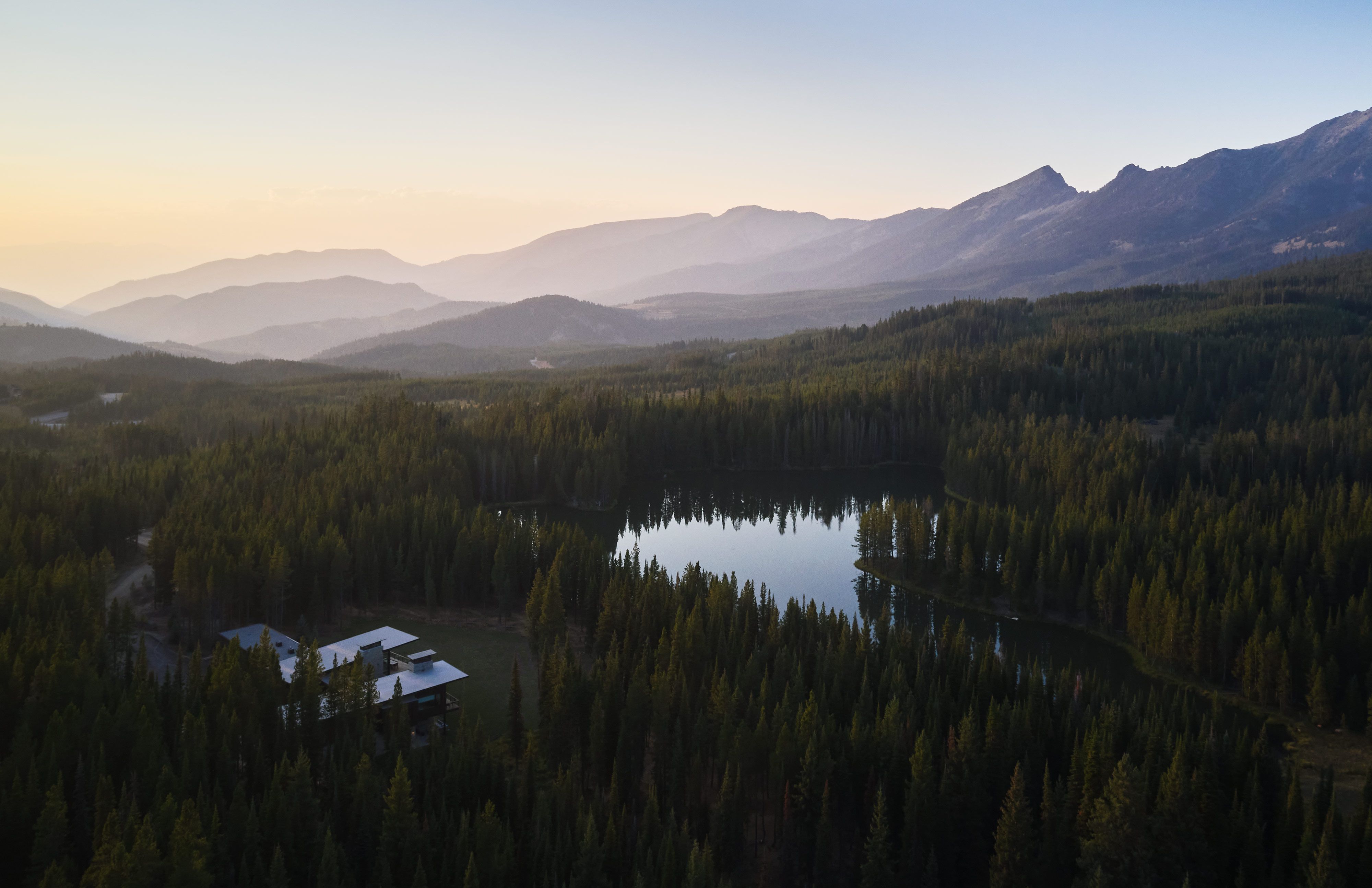 Aerial view of a serene mountain lake reflecting the soft light of sunrise, surrounded by a dense evergreen forest. A modern architectural home is nestled discreetly among the trees, with layers of hazy mountain ranges visible in the distant background.