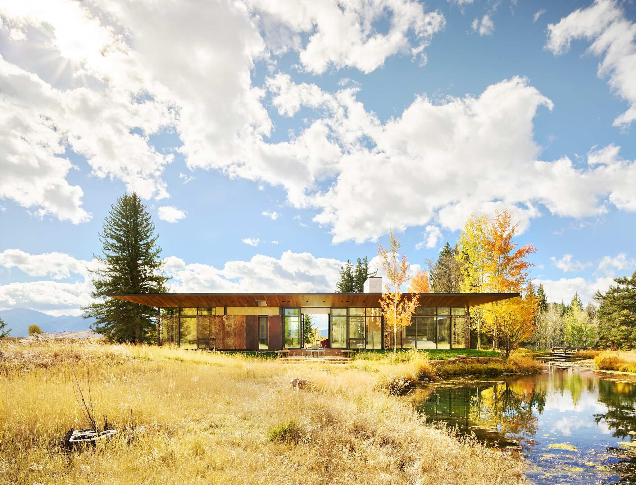 Contemporary glass-walled house with an overhanging roof, set in a golden autumn field beside a reflective pond, with distant mountains under a bright, cloudy blue sky.