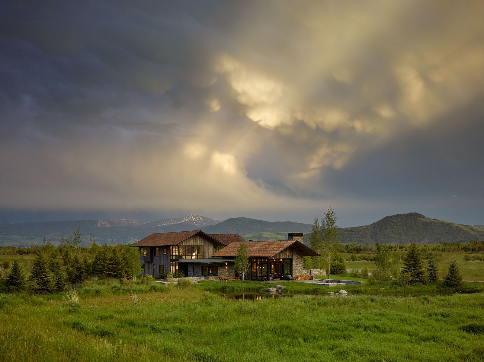 Distant landscape view of a contemporary stone and wood home nestled in a sprawling green field under a dramatic, cloudy sky with sunbeams piercing through over the mountains.