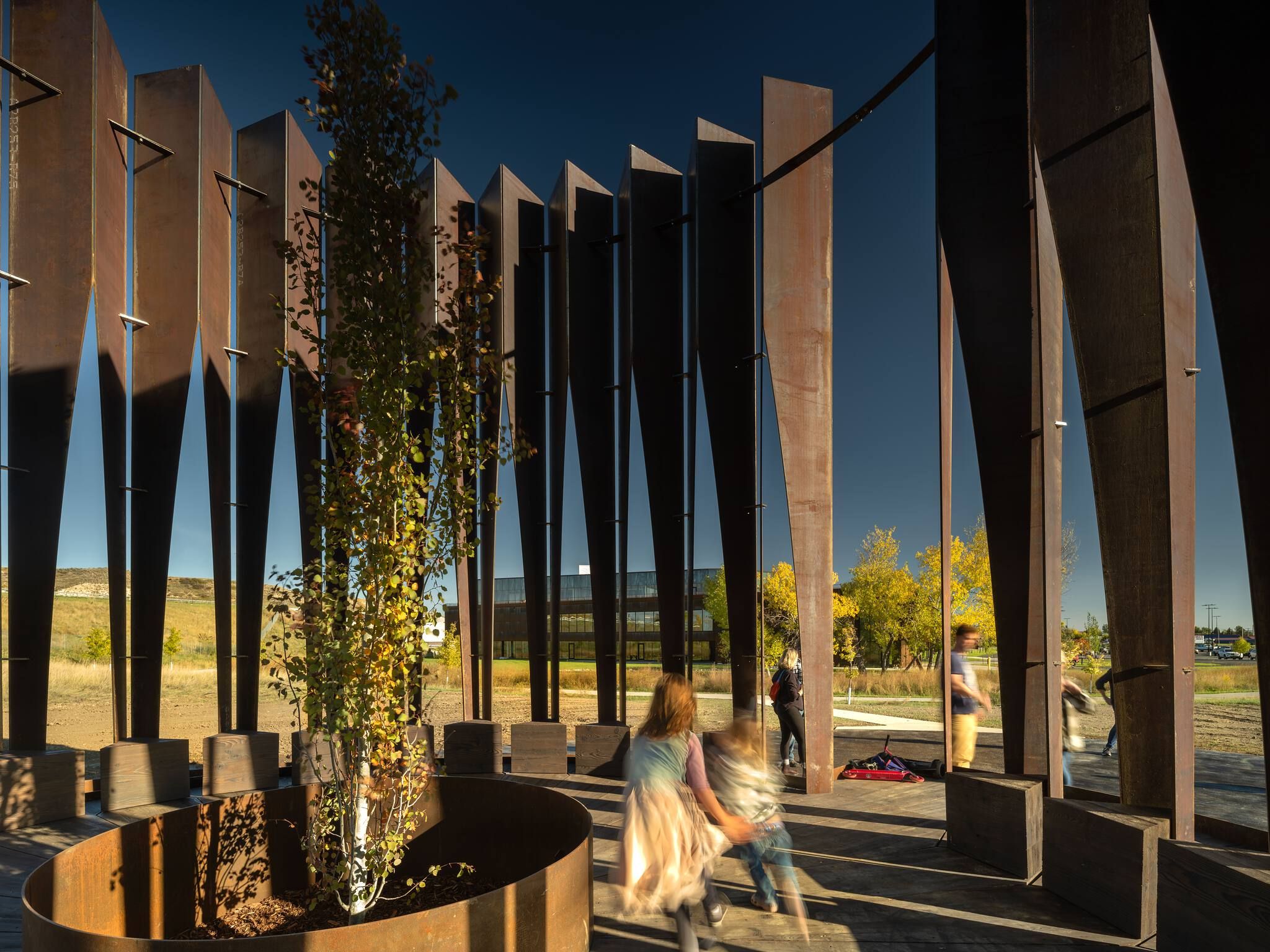 A modern outdoor pavilion made of tall, tapered Corten steel panels arranged in a curve, with a young tree in a circular steel planter and wooden seating blocks on a deck. Blurred children play on the deck while other figures move in the background. Beyond the open panels, a sunny autumn landscape features golden trees, a modern glass building, and rolling hills under a blue sky.