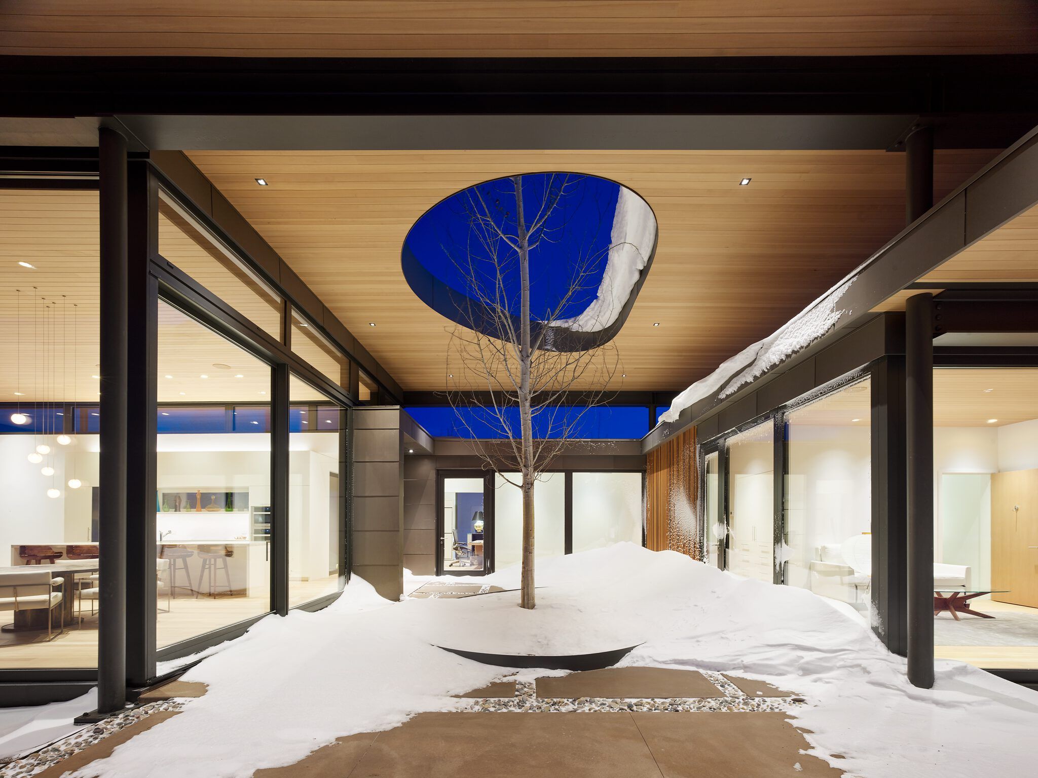 Modern interior courtyard in winter, featuring a central bare tree surrounded by snow and concrete pathways. An irregular oval opening in the wooden ceiling reveals a twilight blue sky, while warm interior lights from surrounding rooms shine through expansive glass walls.