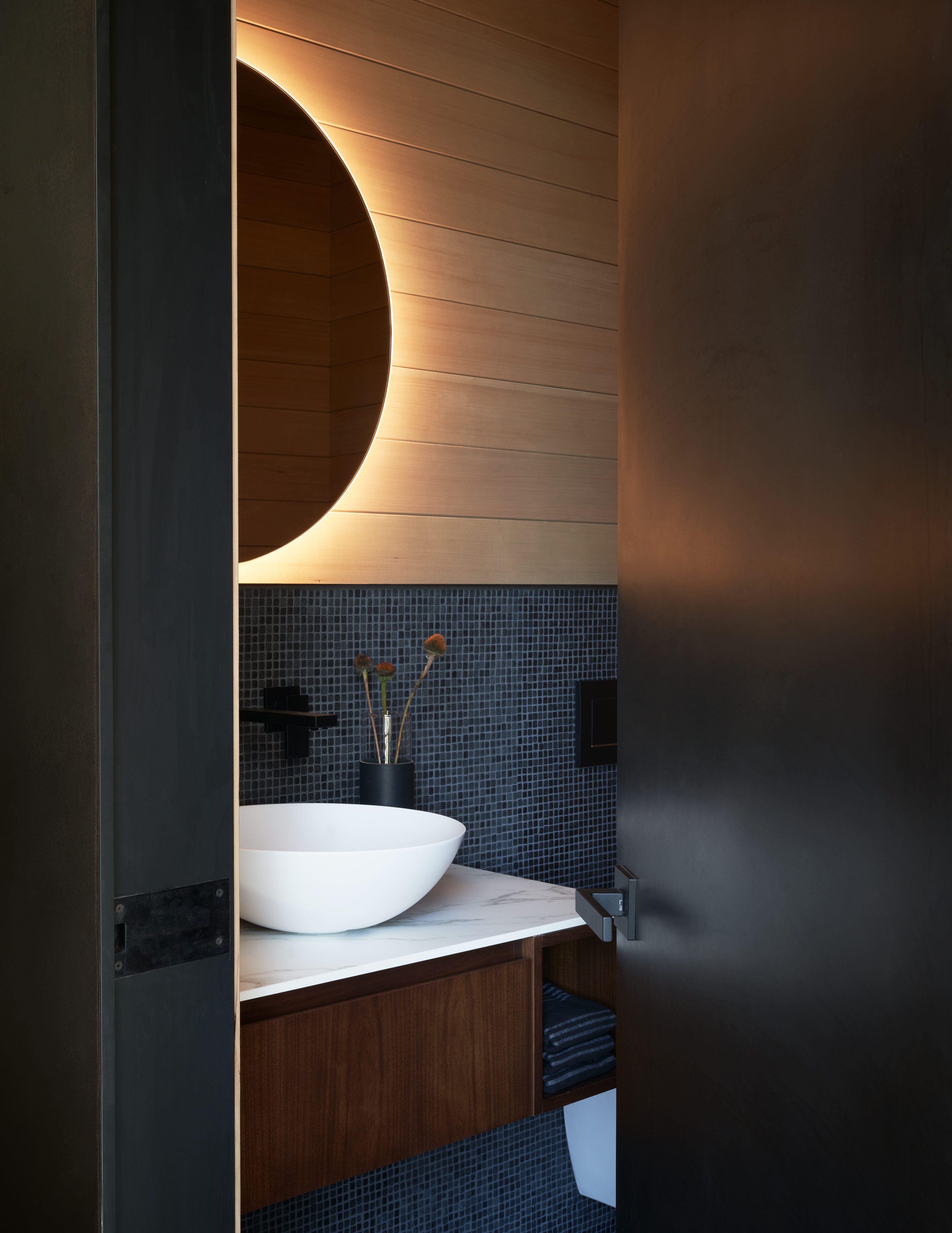 Modern bathroom vanity with a round backlit mirror, light wood paneling, and dark mosaic wall tiles. A white vessel sink sits on a marble countertop above a floating dark wood cabinet, complemented by black fixtures and a subtle vase with dried flowers.