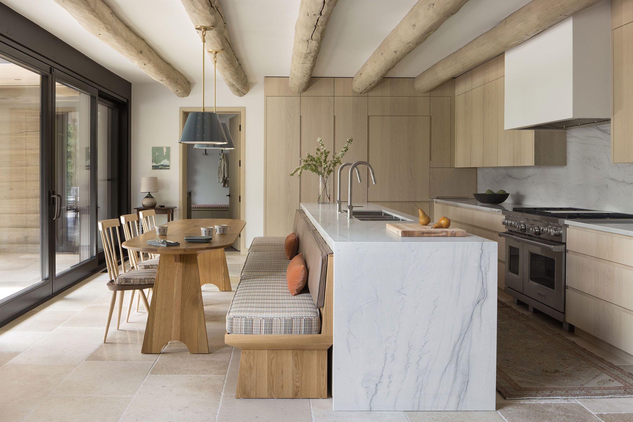 Modern kitchen with light wood cabinetry, exposed ceiling beams, and a large white marble island featuring built-in bench seating facing a matching wooden dining table.