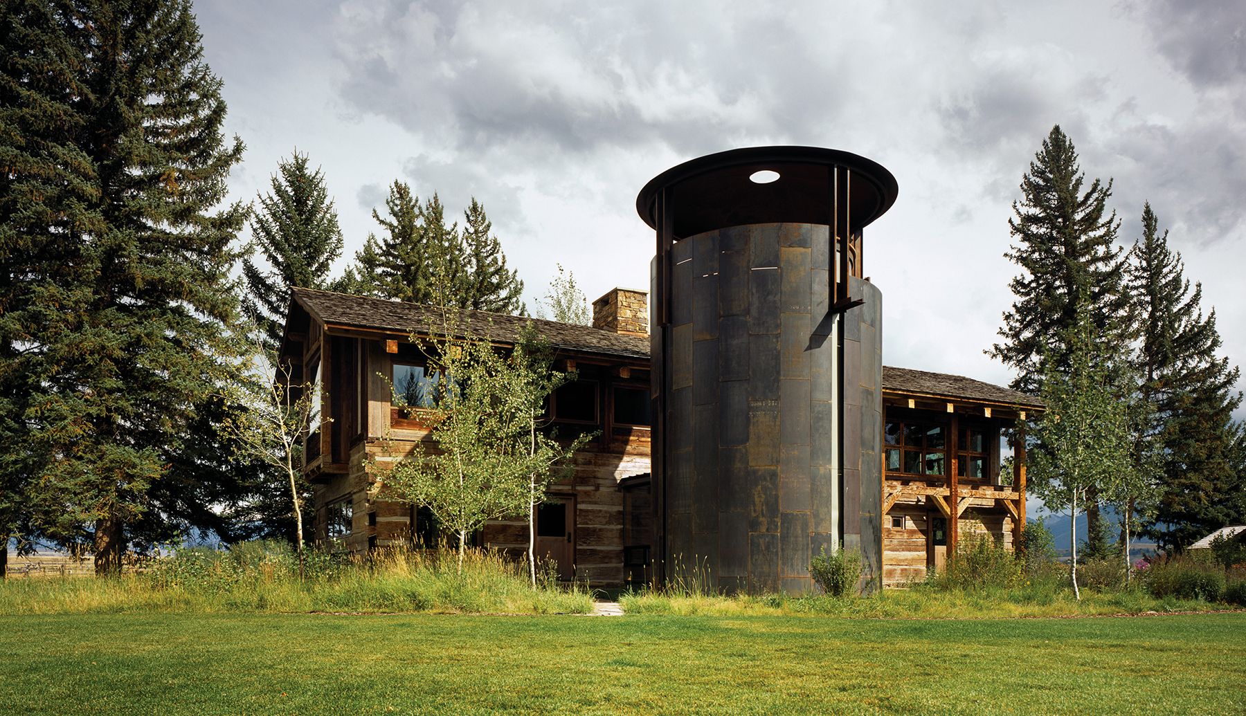 Exterior view of a rustic wooden home with a tall, cylindrical dark metal wine silo structure set against a grassy yard and pine trees.