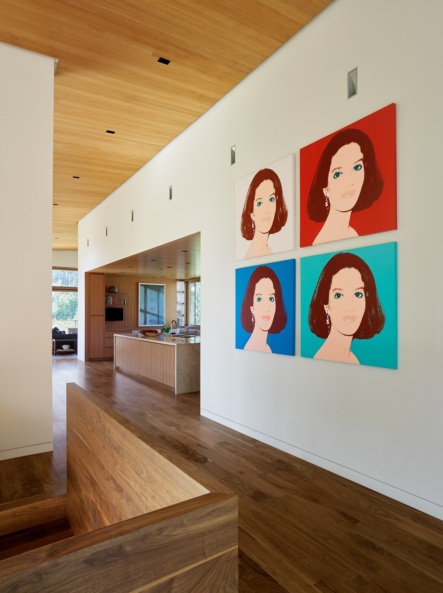 Modern interior hallway with a light wood slat ceiling and dark wood floors, featuring a long white wall adorned with four colorful Pop Art portraits of a woman. The hallway leads to an open-concept kitchen and dining area with wood cabinetry, a large island, and natural light from large windows. A wood stair railing is visible in the foreground.