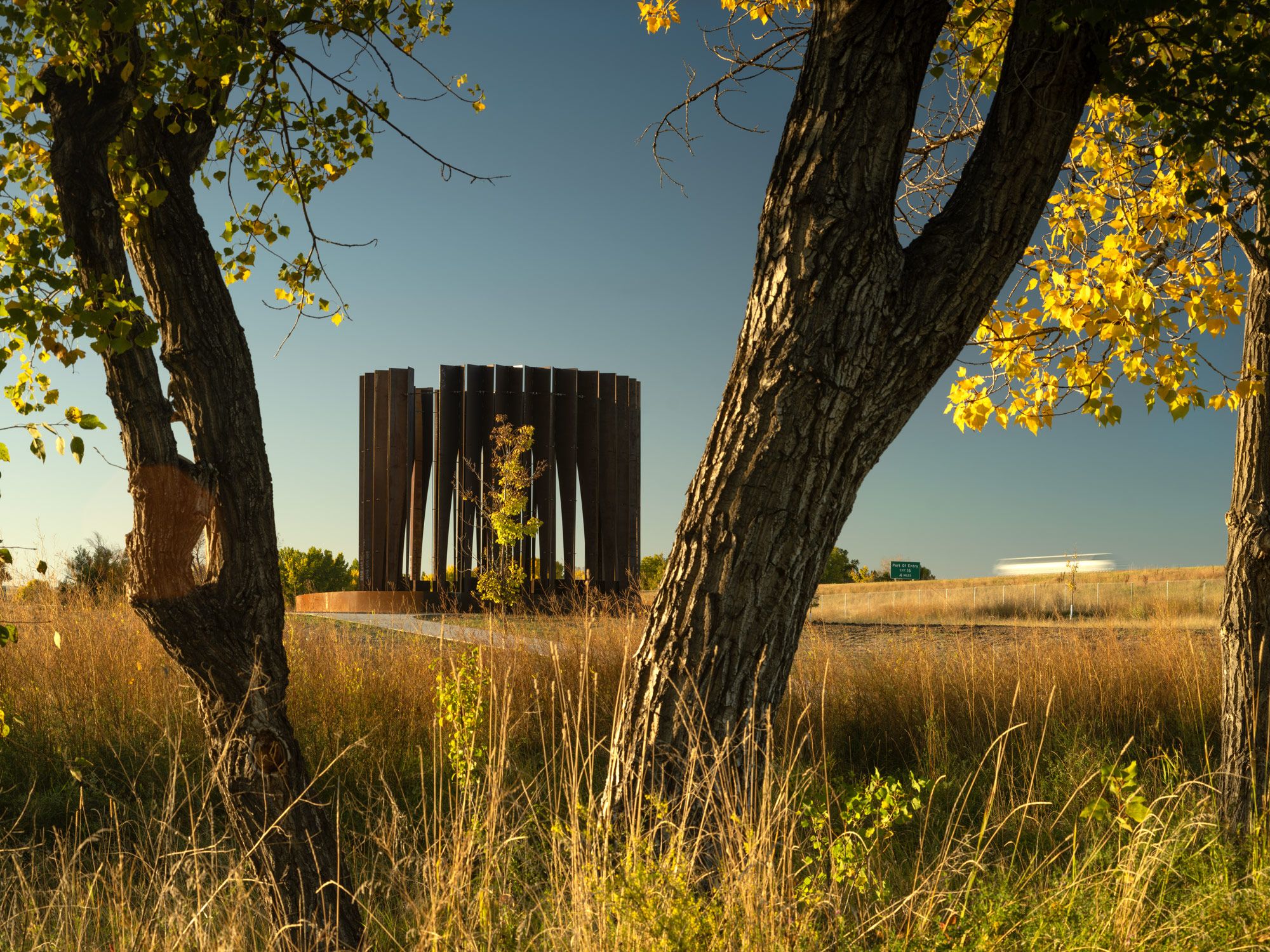 Dark, vertical-paneled circular monument set in a golden prairie, framed by two gnarled trees with vibrant yellow autumn leaves. A highway with a streaking vehicle and a distant green sign is visible under a clear blue sky.
