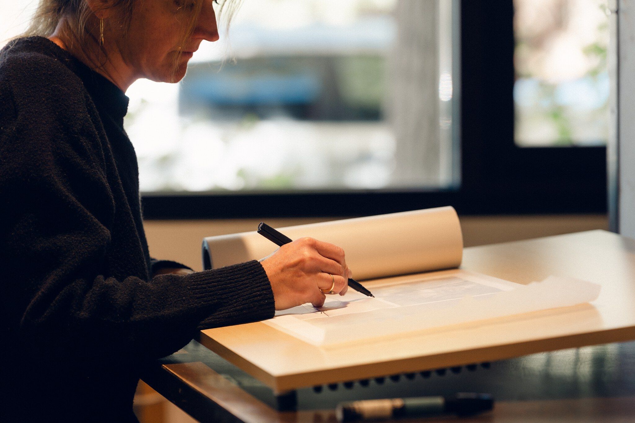 Side profile of a woman with light hair, wearing a dark textured sweater, intently drawing on a sheet of paper with a black pen. A gold ring adorns her finger as she works at a light wooden desk, with a roll of paper and a blurred bright window visible in the background.