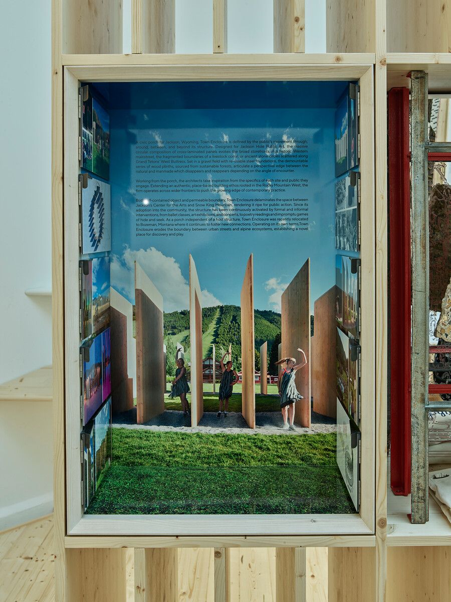 Exhibition display showing the Town Enclosure public art installation, featuring tall wooden panels with young girls playing among them on a grassy landscape, set against a mountain ski slope and blue sky. Accompanying text describes the project's purpose and its original location in Jackson, Wyoming.