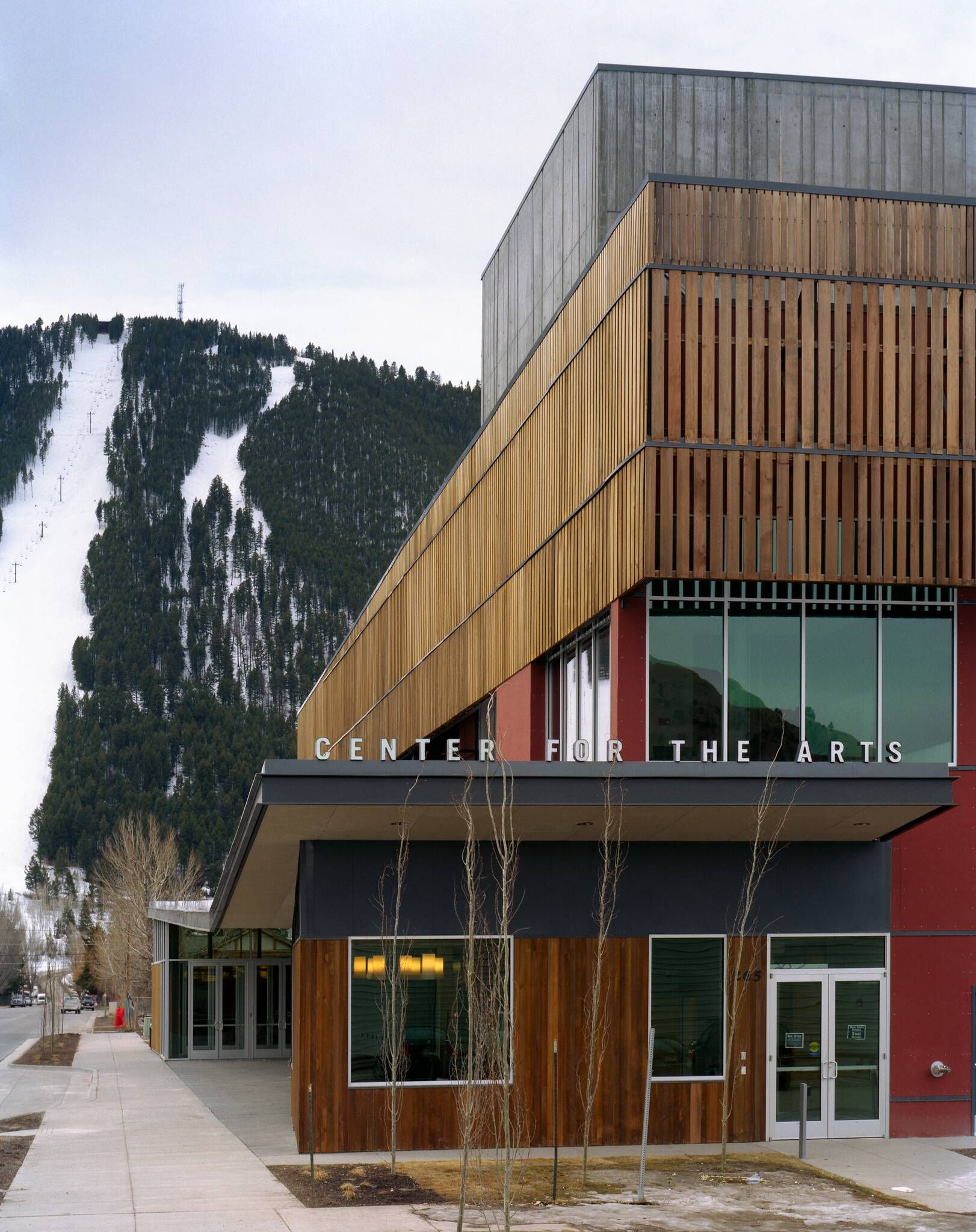 Modern Center for the Arts building with a red, concrete, and slatted wood facade, featuring CENTER FOR THE ARTS signage, set against a snow-covered mountain with ski slopes and pine trees.