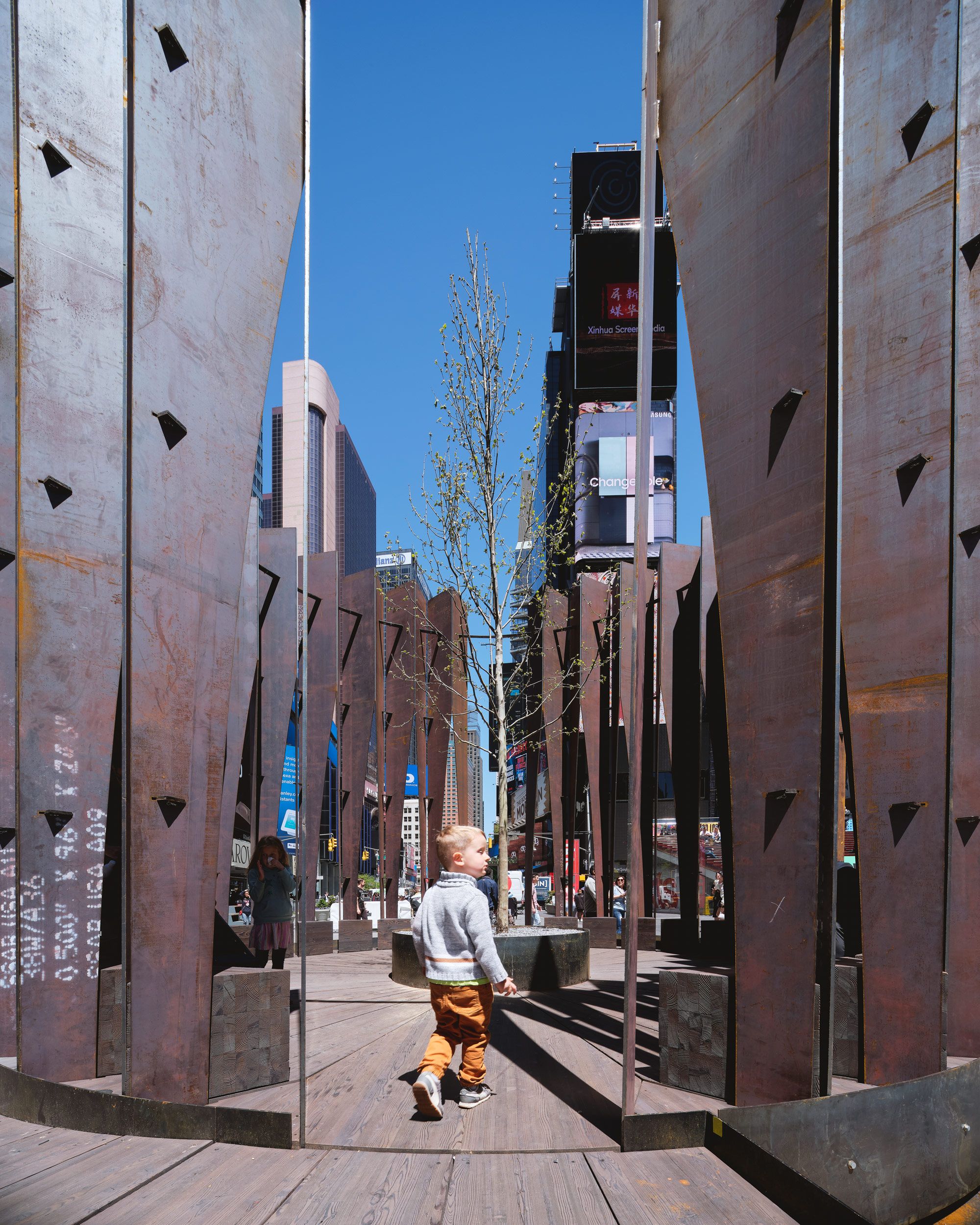 A young boy walks on a wooden path amidst an urban art installation featuring tall, weathered metal panels. Mirrored panels reflect the boy, surrounding city buildings, and bright digital billboards under a clear blue sky.