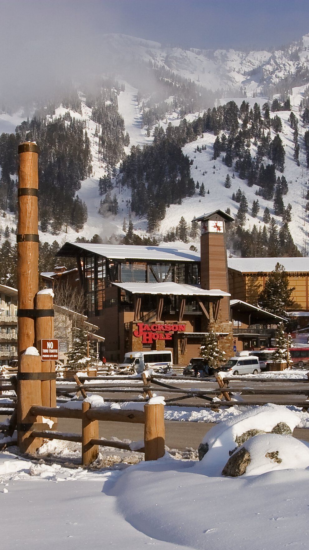 Jackson Hole Mountain Resort base area in winter, with snow-covered ski slopes and pine trees rising behind modern lodge buildings. The main building displays a red 'Jackson Hole' sign and a tall clock tower, while fresh snow blankets a rustic wooden fence in the foreground.