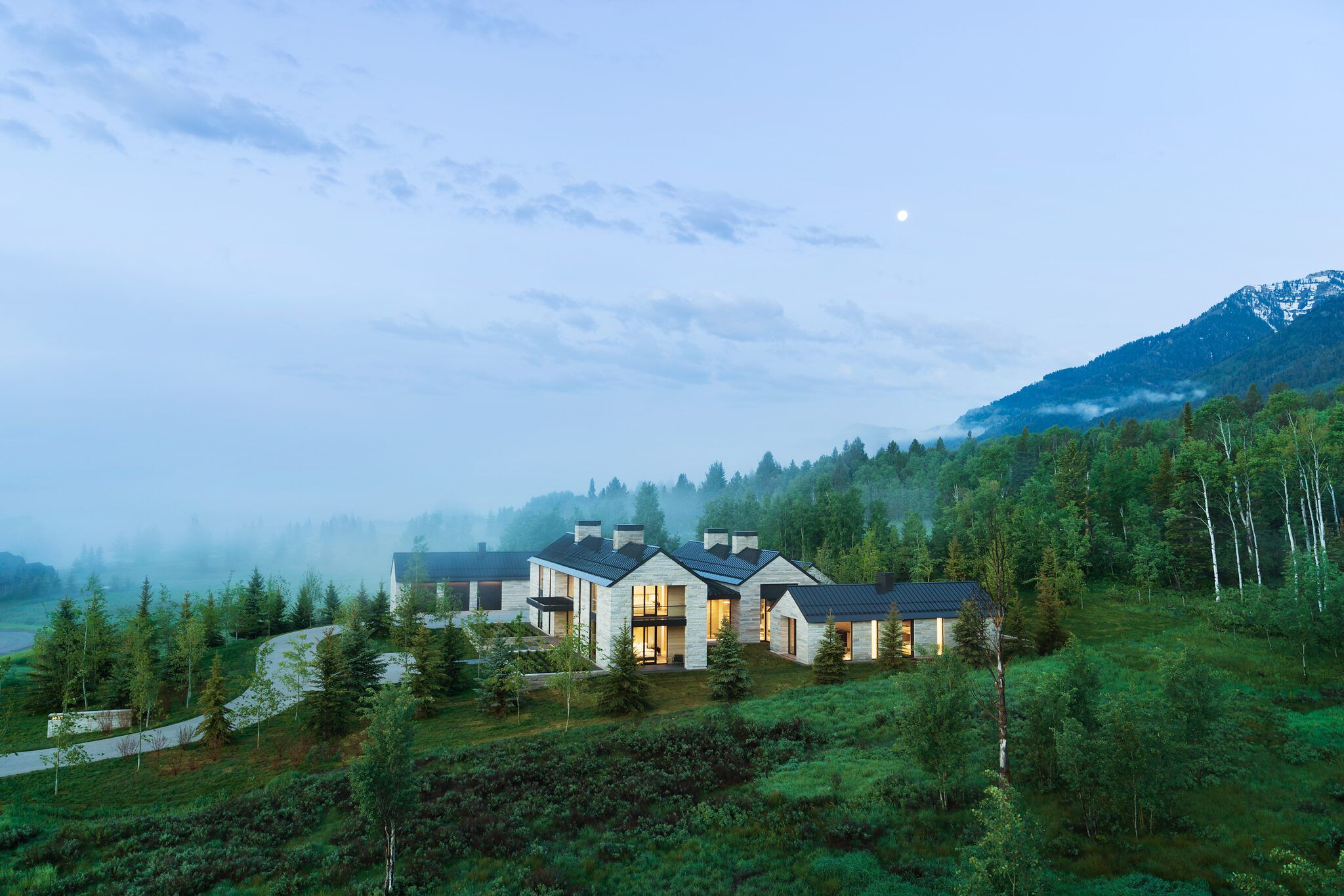 Sprawling contemporary stone mountain house with dark roofs and illuminated windows, set on a lush green hillside. Misty forests and distant mountains are visible under a twilight sky with a prominent moon.