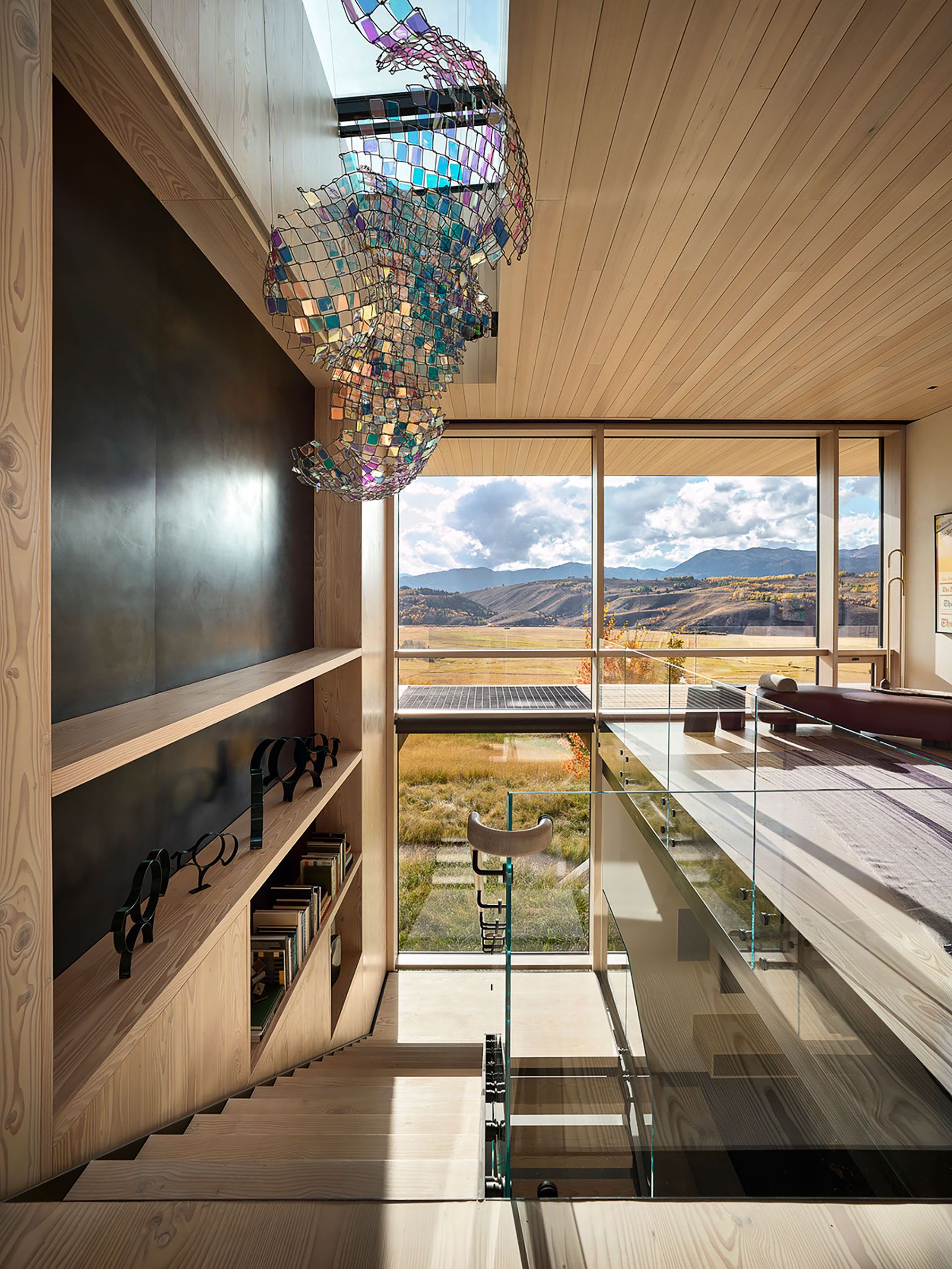 Modern interior wooden staircase and upper loft featuring a colorful glass mesh light fixture and floor-to-ceiling view of distant mountains.