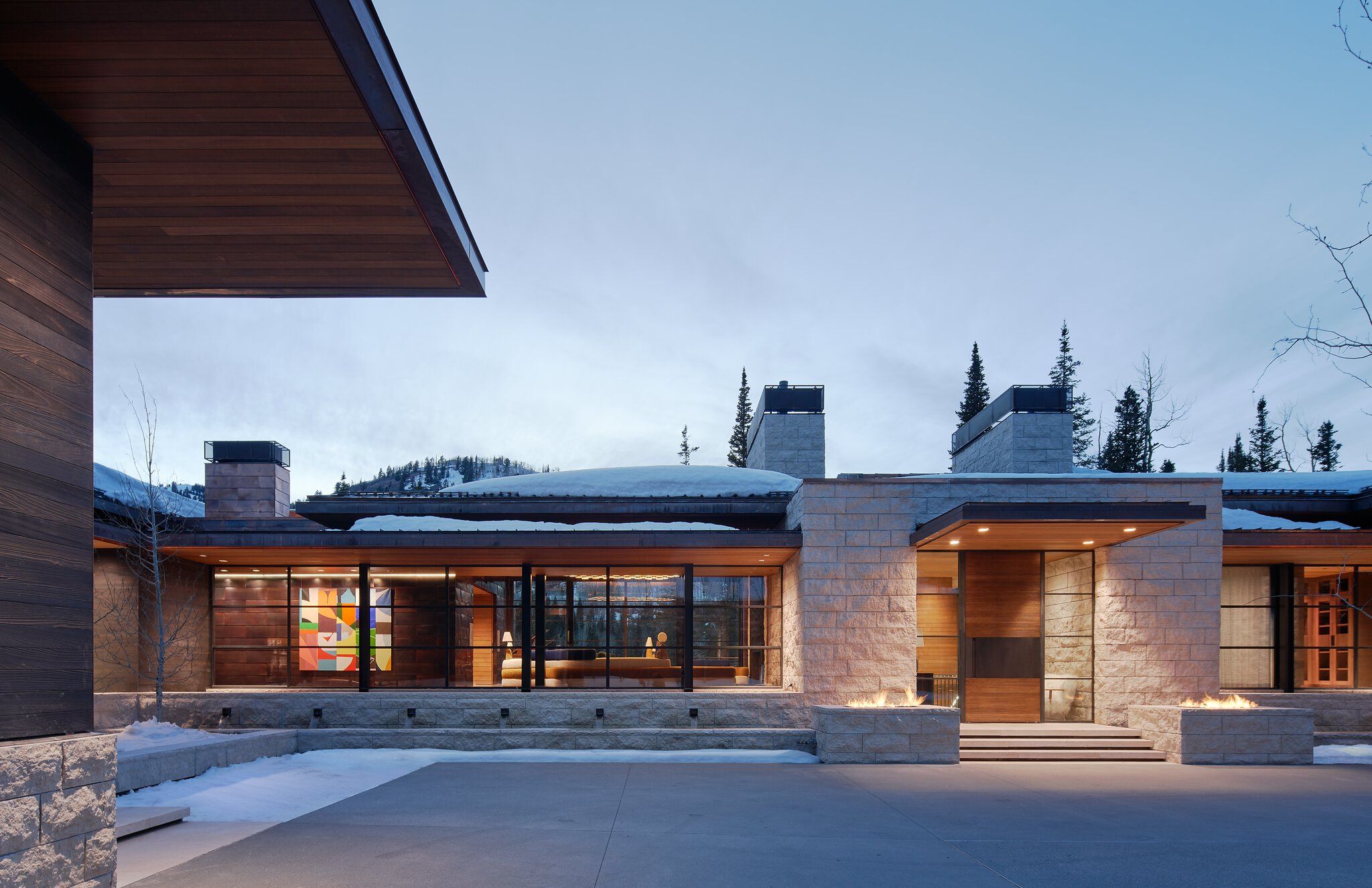 A contemporary mountain home with stone and dark wood exterior, featuring large glass windows, an illuminated entrance, and two outdoor fire pits, set in a snowy landscape at dusk.