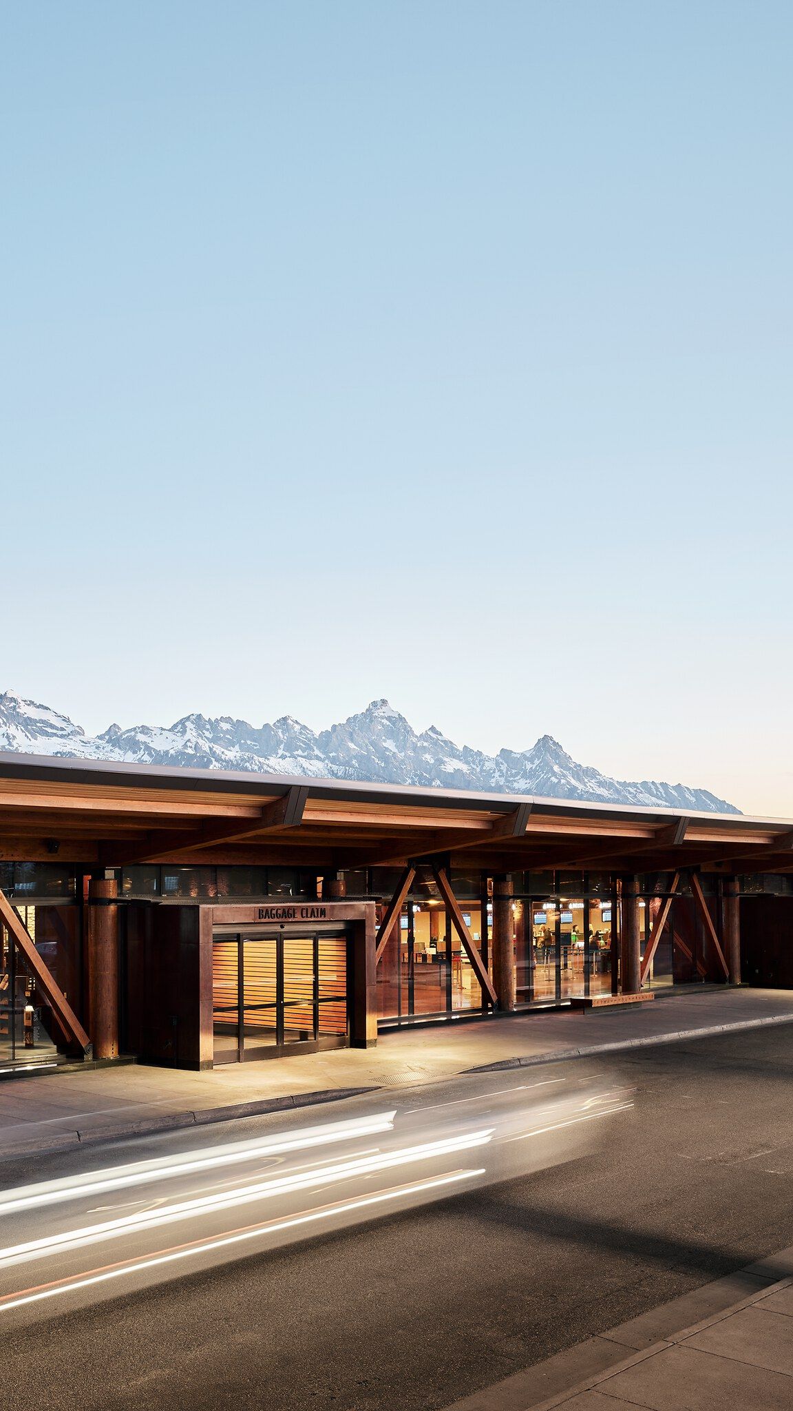 Lodge-style airport terminal at dusk with a prominent BAGGAGE CLAIM sign, glowing interior lights, and snow-capped mountains in the background. Car light trails streak across the road in the foreground.