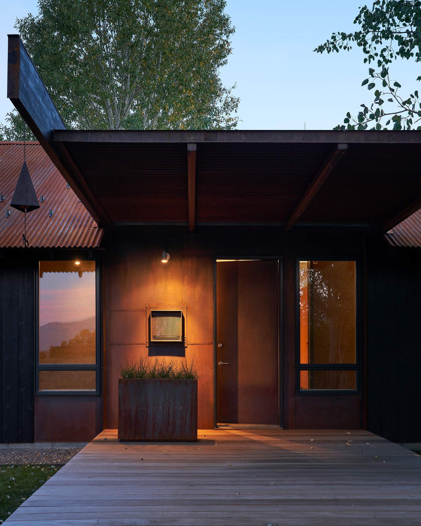 Modern building entrance at dusk, featuring a weathered Corten steel facade and corrugated roof. A light wooden deck leads to the dark door, illuminated by a warm exterior light and interior window glow. A metal planter with greenery sits beside the door, with trees visible against a blue sky.