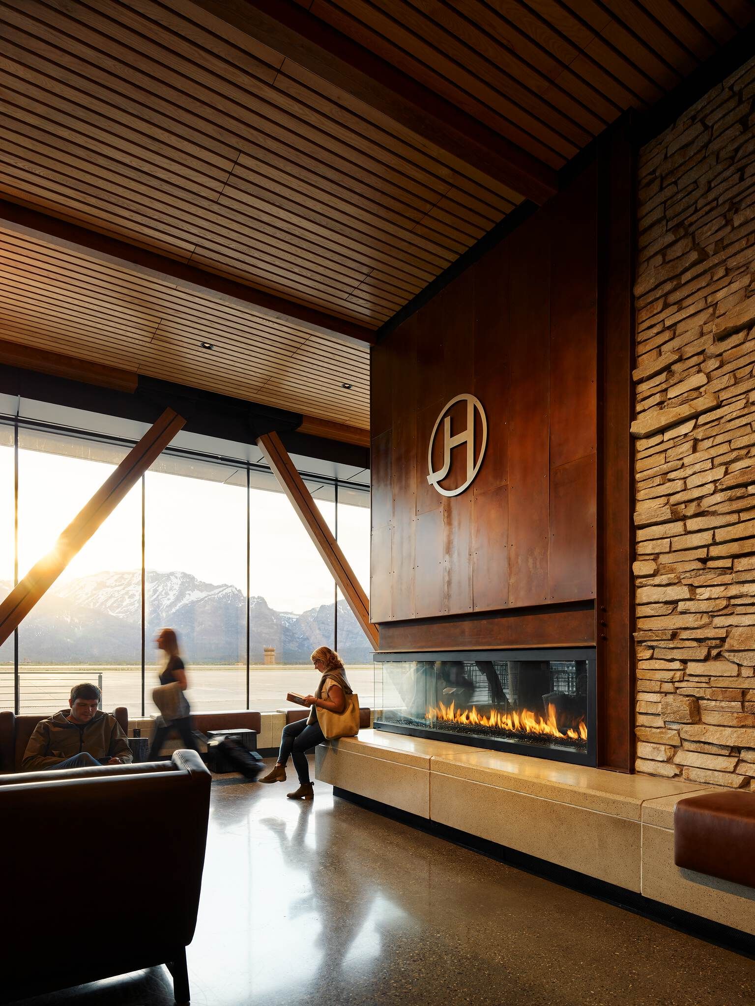 Modern lodge-style airport lounge with a large, horizontal gas fireplace set into a rustic metal wall, featuring a circular logo. Adjacent stone wall and wood-slatted ceiling. Floor-to-ceiling windows offer sunset views of snow-capped mountains and an airport tarmac with travelers relaxing in lounge chairs and a woman reading on a bench.