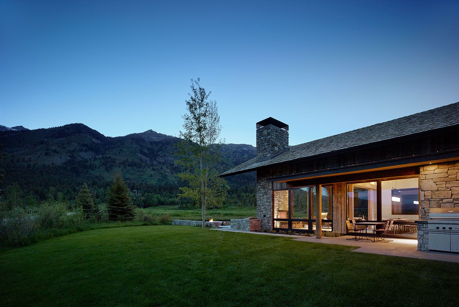 Wide exterior shot at twilight showing the back patio of a modern rustic home with stone accents, a built-in grill, and a smooth lawn stretching toward dark mountain silhouettes.
