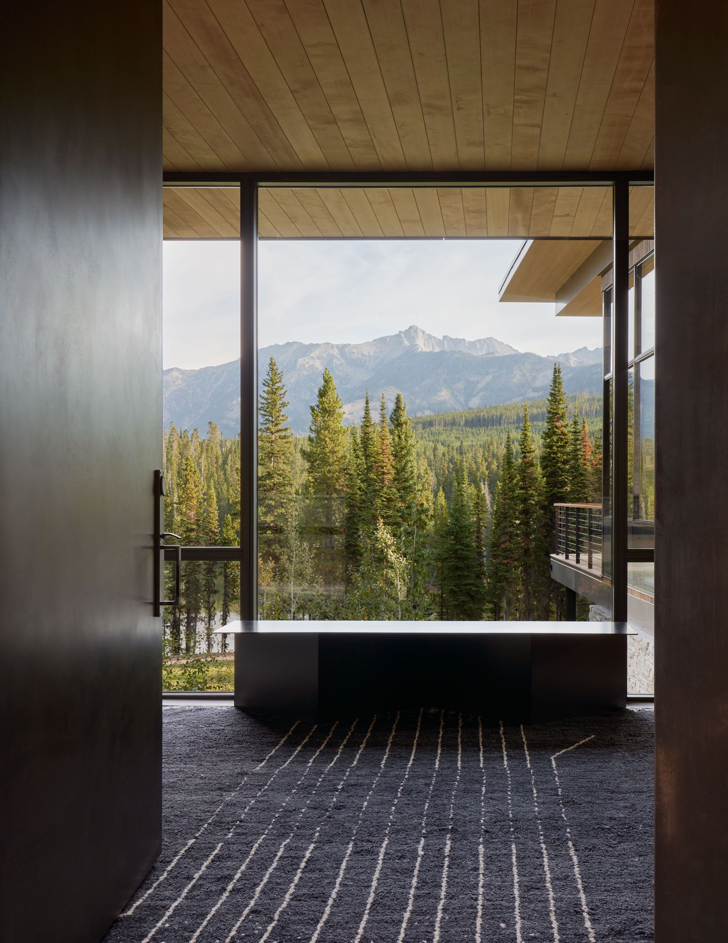 Panoramic view from a modern interior featuring a dense pine forest and majestic, rugged mountains under a bright sky. The room includes a dark, textured rug with light vertical stripes, a minimalist dark console table by the expansive window, and a light wood-slat ceiling, framed by a dark wall on the left.