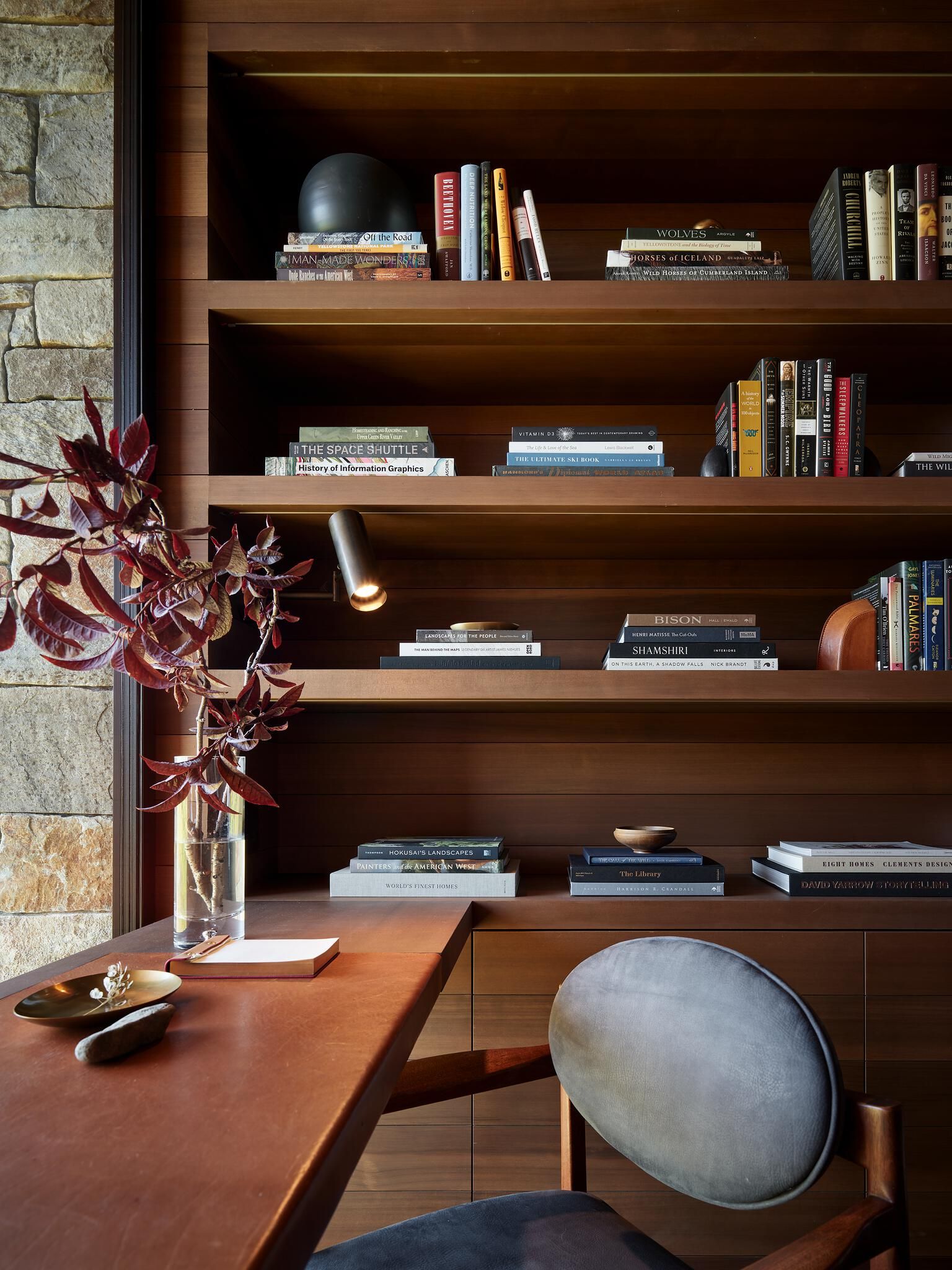Modern built-in wooden bookshelves and desk in a sophisticated home office or study, featuring various books, decorative objects, and a sleek spotlight. A vase of red foliage, an open notebook, and a brass dish adorn the desk. A grey upholstered chair and a rustic stone wall complete the cozy space.