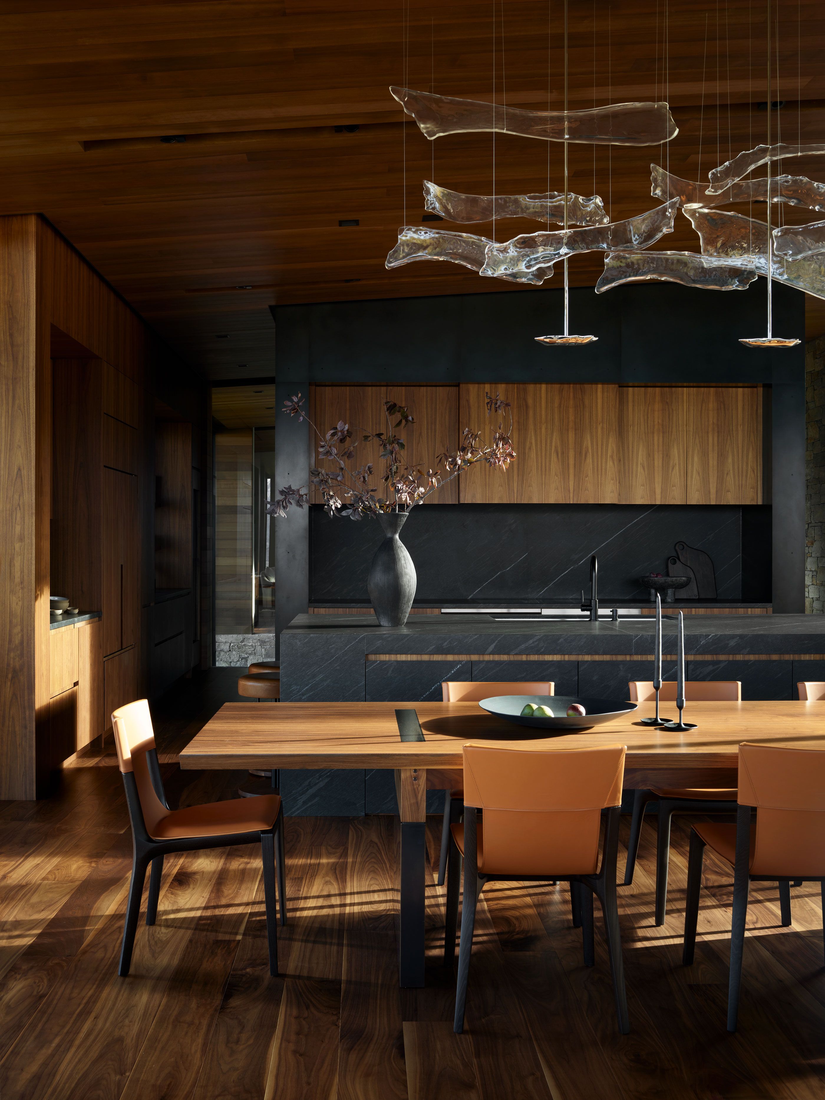 Modern open-plan kitchen and dining room featuring a long wooden table with tan leather chairs and an artistic multi-paneled glass chandelier. The kitchen area showcases dark stone countertops, backsplash, and island, complemented by warm wood upper cabinetry and a dark wood floor and ceiling.