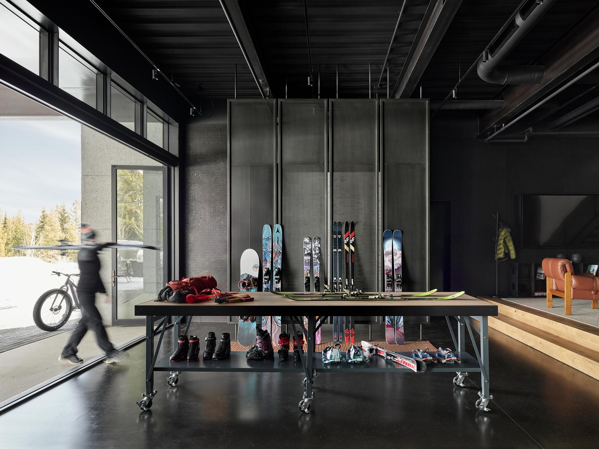 Modern industrial ski gear room with a long dark table displaying multiple pairs of skis, snowboards, and boots. A blurred figure carries skis out through a large sliding glass door to a snowy landscape, while a raised living area with a brown leather armchair is visible in the background.