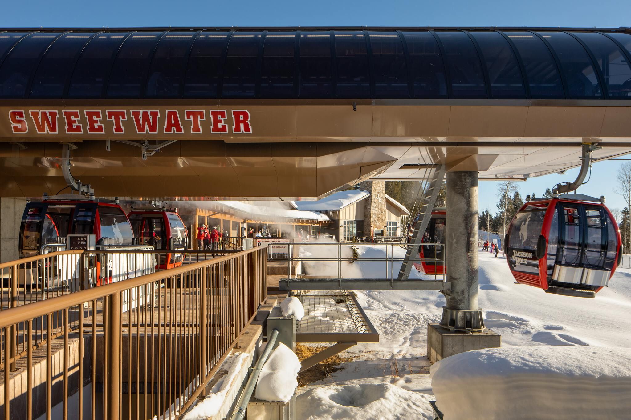 The Sweetwater gondola station at Jackson Hole Mountain Resort, showing red gondola cabins at the loading platform with people, and one cabin traveling on the right over a snowy landscape under a clear blue sky. A rustic building is visible in the background.