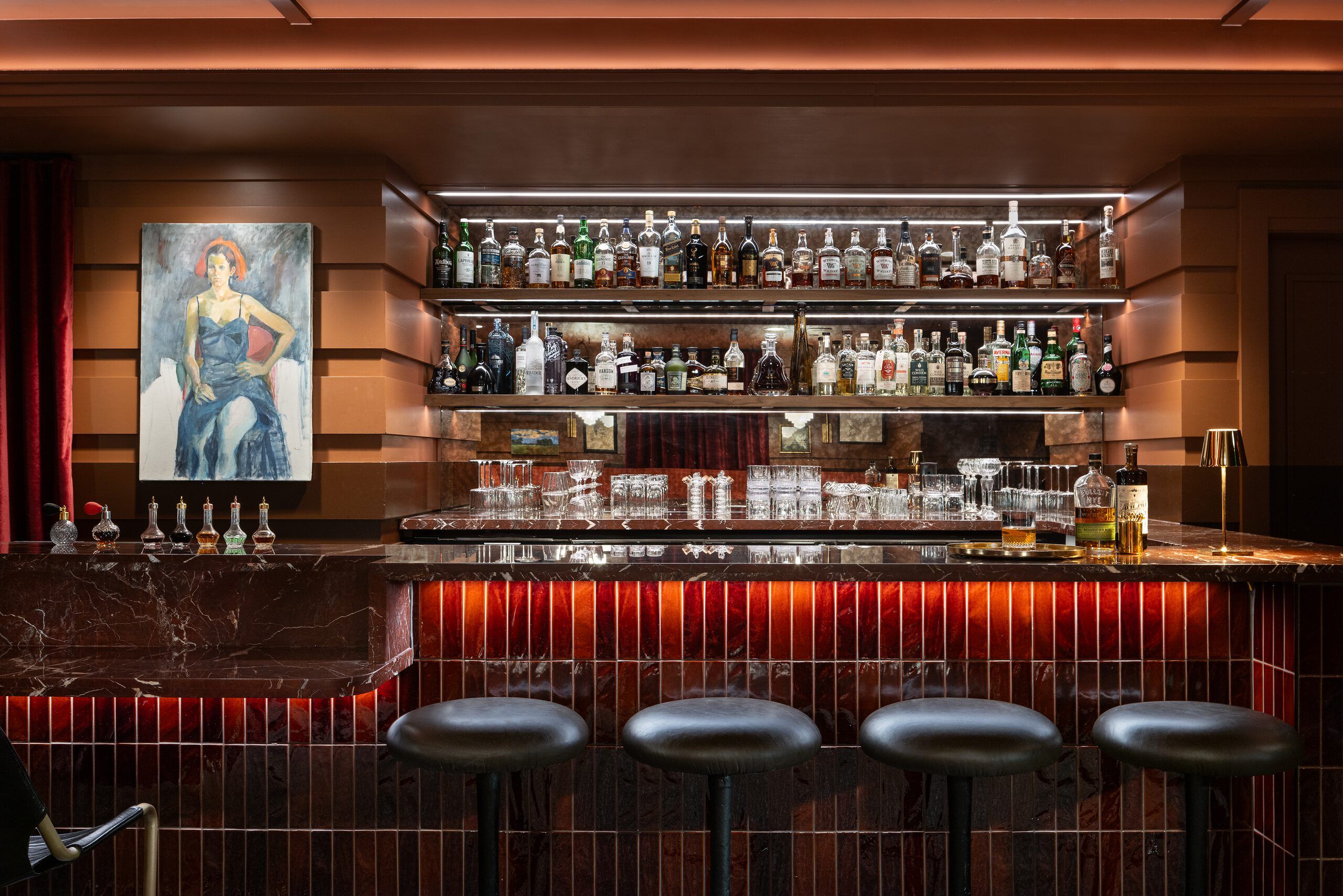 Elegant bar interior with illuminated liquor shelves, a dark marble counter featuring glowing red-orange vertical tiles, and four black bar stools. A painting of a woman hangs on the wall to the left.
