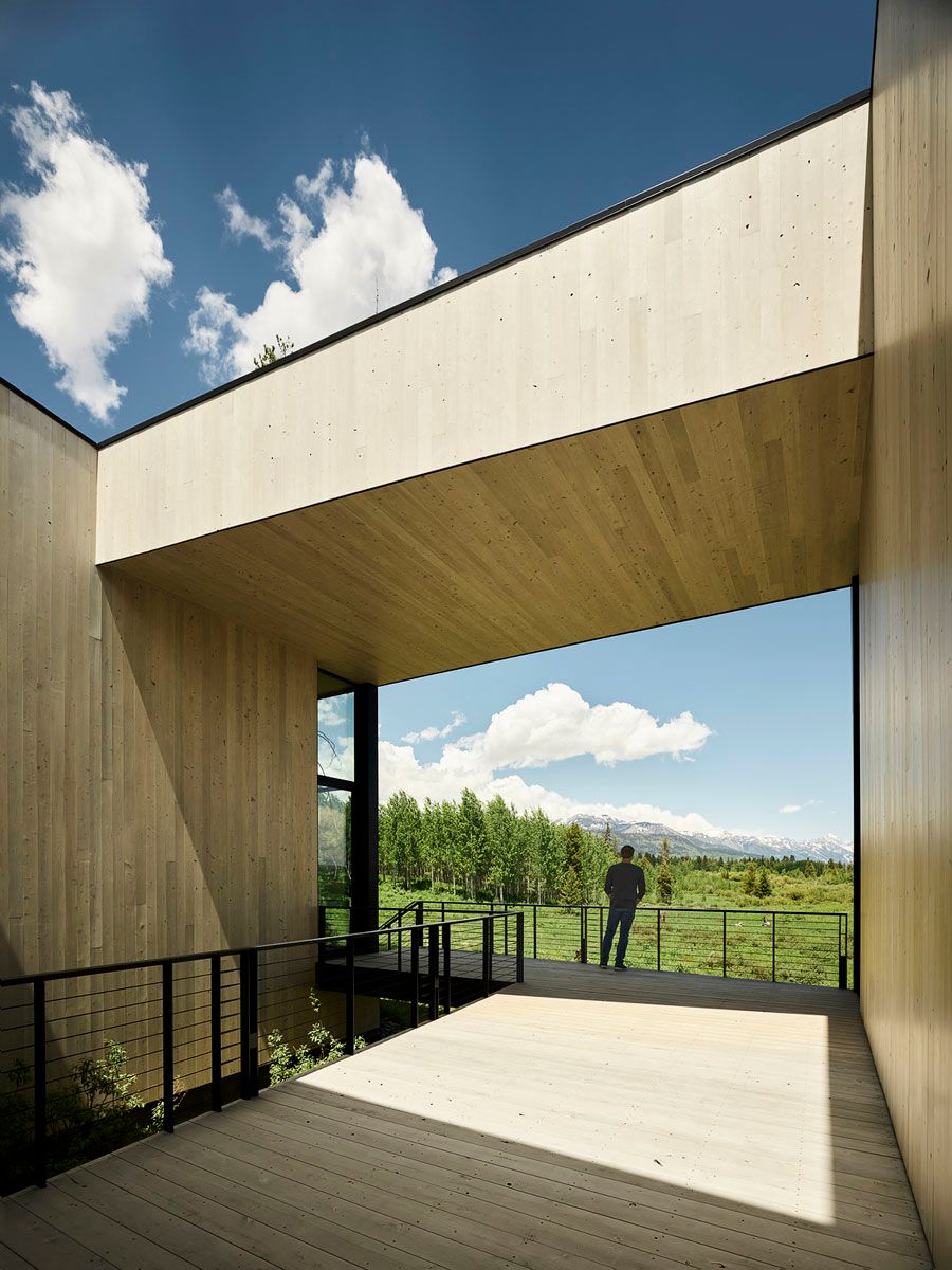 A person stands on a modern wooden deck, part of a contemporary building with light wood paneling, observing a panoramic vista of green meadows, an aspen forest, and distant snow-capped mountains under a blue sky with white clouds.