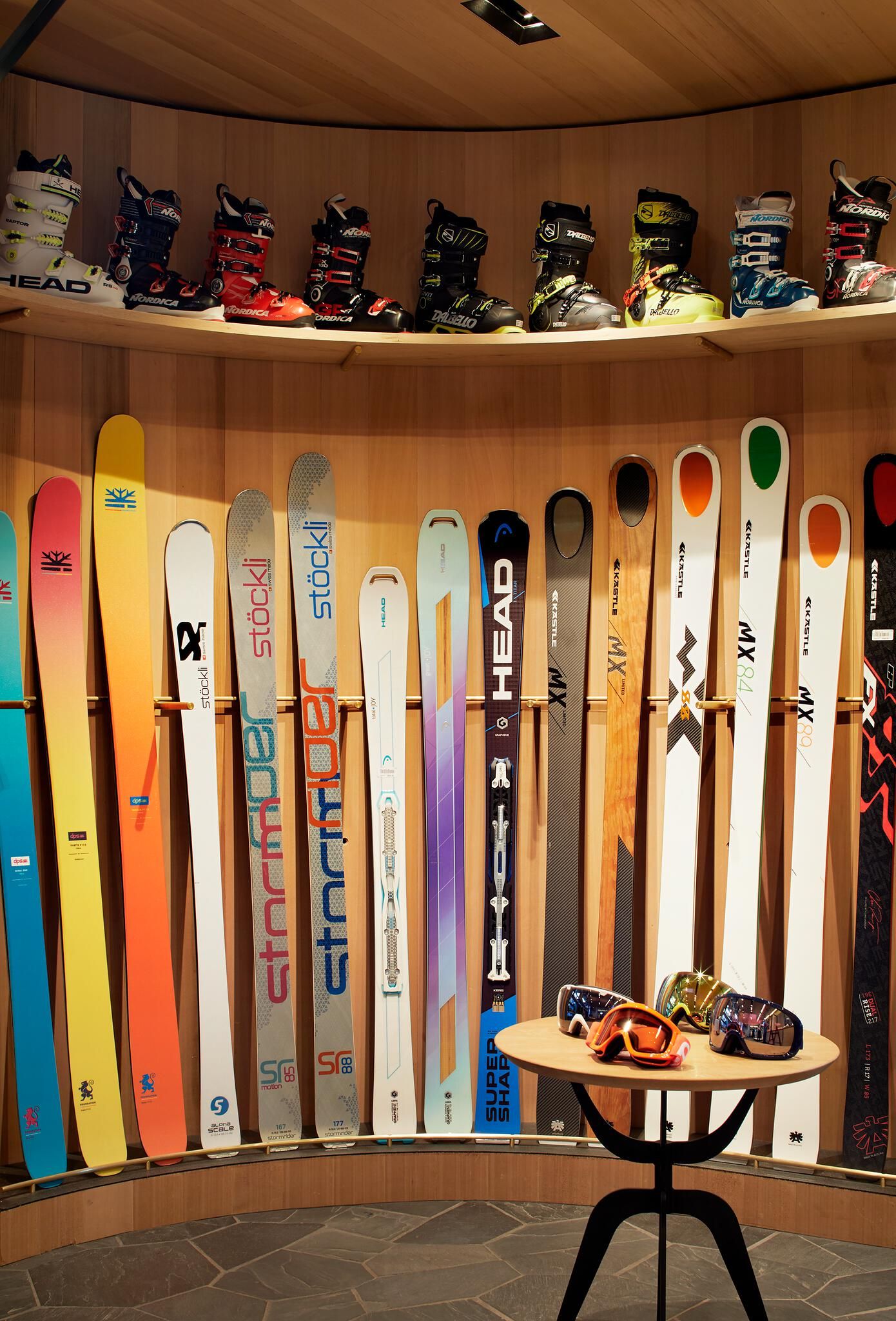 A comprehensive display of ski equipment featuring multiple pairs of ski boots from brands like HEAD, Nordica, and Dalbello on two upper curved wooden shelves. Below, a long row of various skis stand upright with diverse colors and graphics. In the foreground, four pairs of colorful ski goggles are arranged on a small round wooden table in a well-lit ski shop setting.