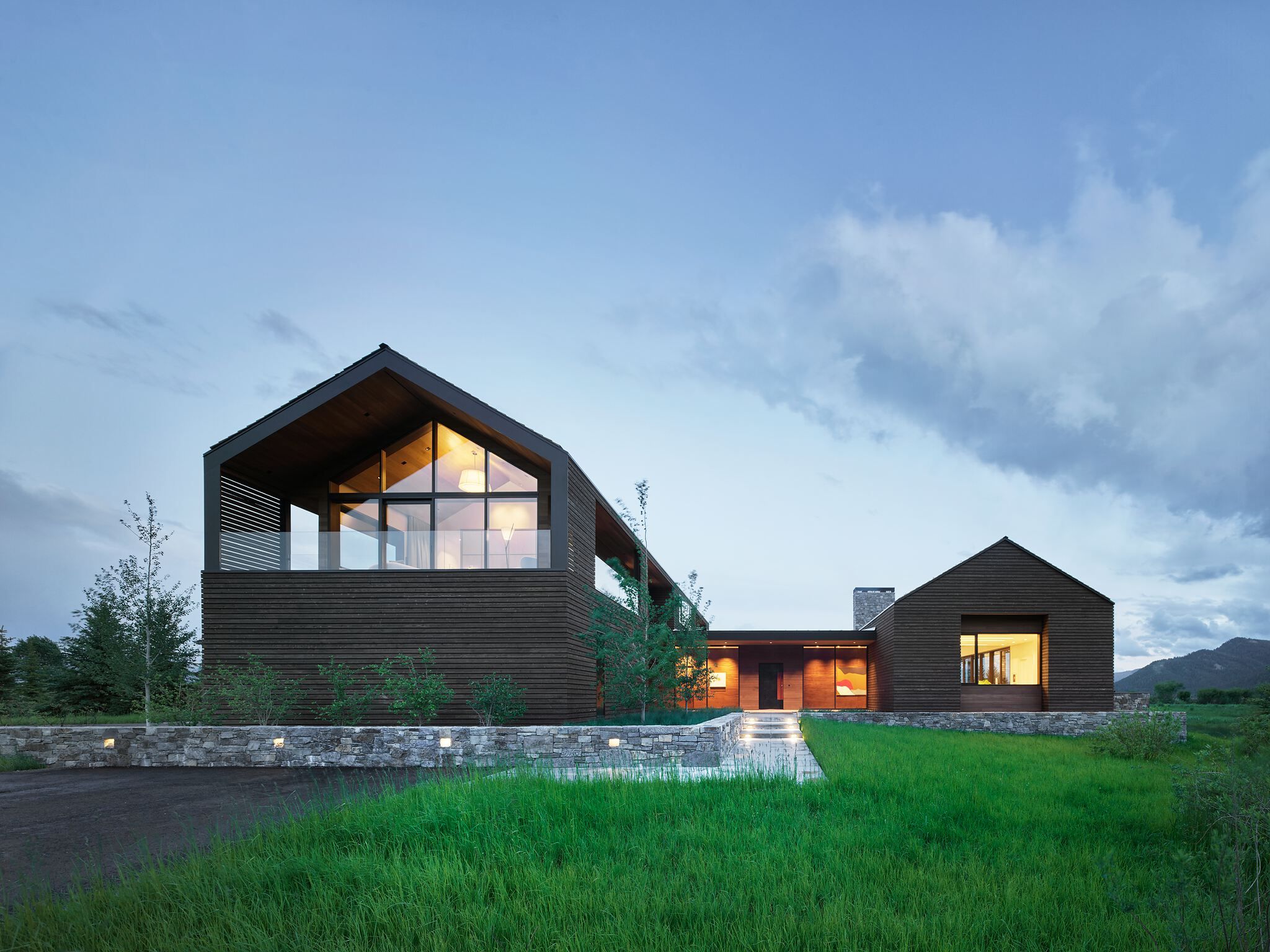 Modern dark-clad luxury home at dusk, featuring gabled architecture, large windows, an illuminated stone wall, and a glowing pathway leading through green grass with mountains in the background.
