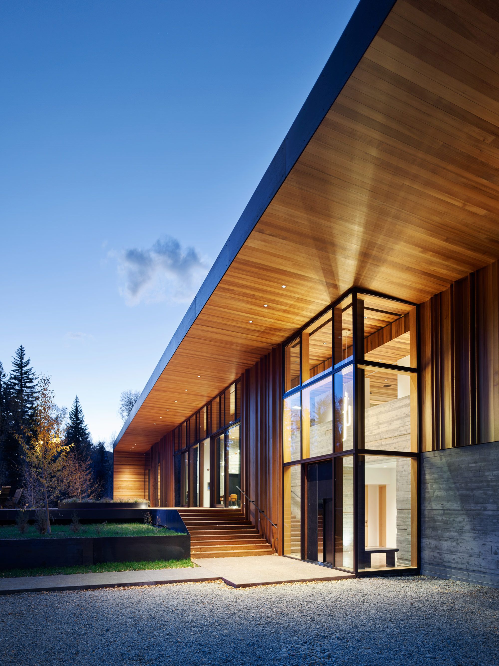 Contemporary building exterior at dusk, featuring a large wooden overhanging roof, floor-to-ceiling glass windows, wood paneling, and concrete elements, with warm lights illuminating the interior and exterior, a gravel foreground, and trees in the background.