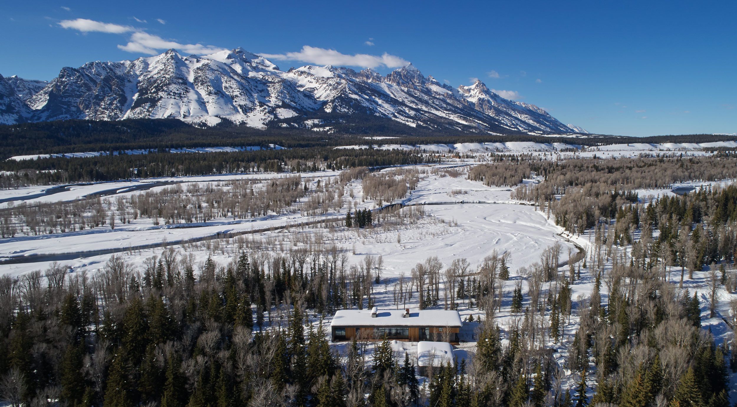 Aerial view of a modern house with a snow-covered roof nestled in a winter forest. Beyond, a winding river flows through a snow-laden valley, with the majestic, snow-capped Grand Teton mountains rising under a clear blue sky.