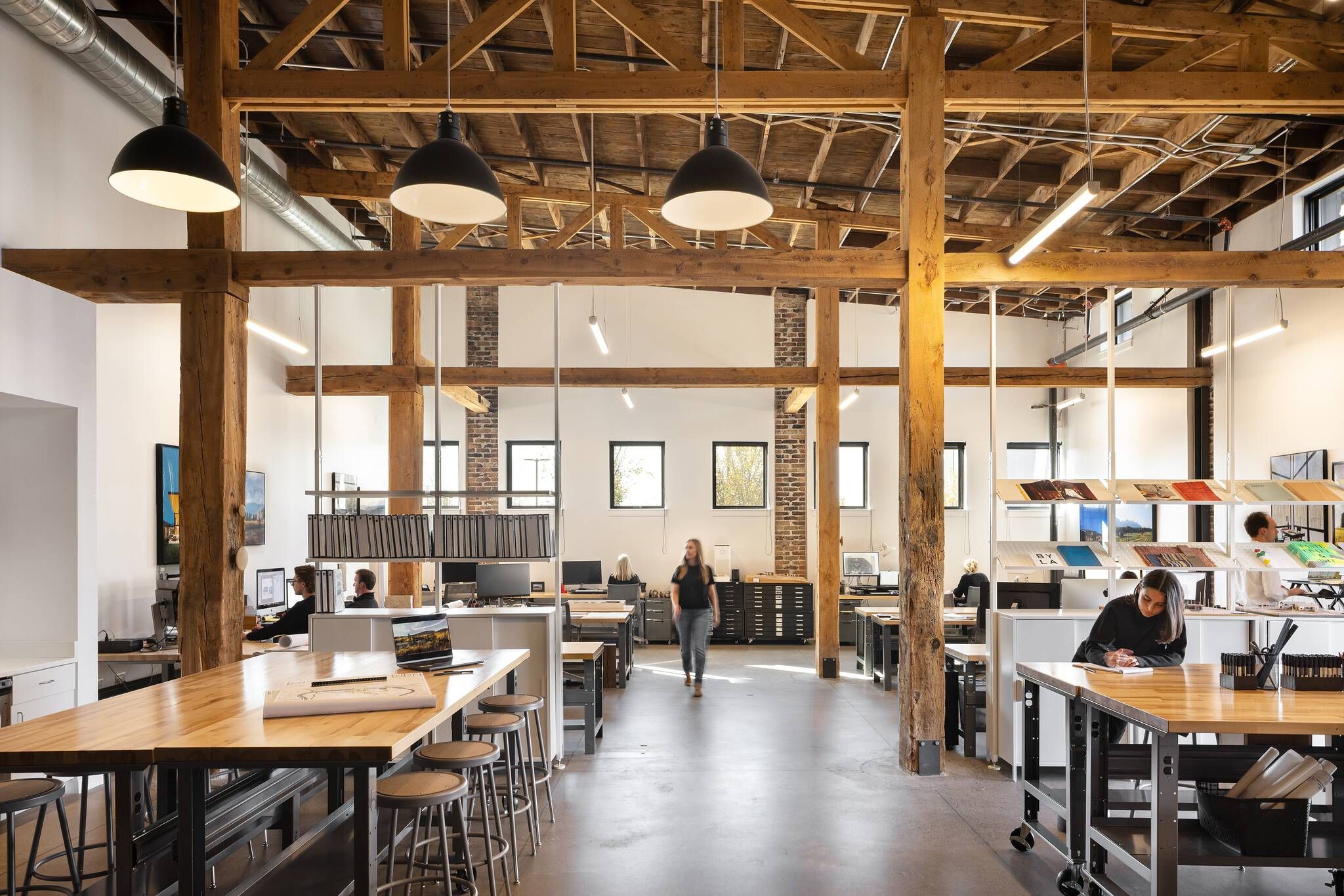Modern open-plan office interior featuring exposed timber beams and columns, high ceilings, and polished concrete floors. Employees work at individual desks and a long communal wooden table with stools. Industrial pendant lights, linear fixtures, and large windows illuminate the collaborative workspace, which includes exposed brick accents and architectural sample displays.