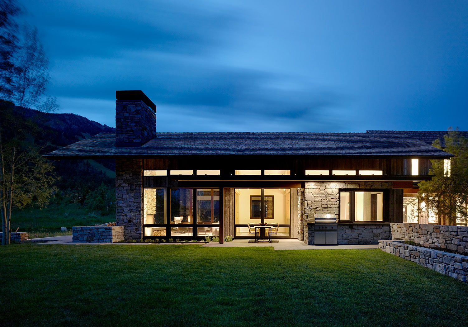 Exterior dusk shot of a modern mountain home with illuminated interiors, a stone chimney, and a manicured green lawn under a deep blue twilight sky.