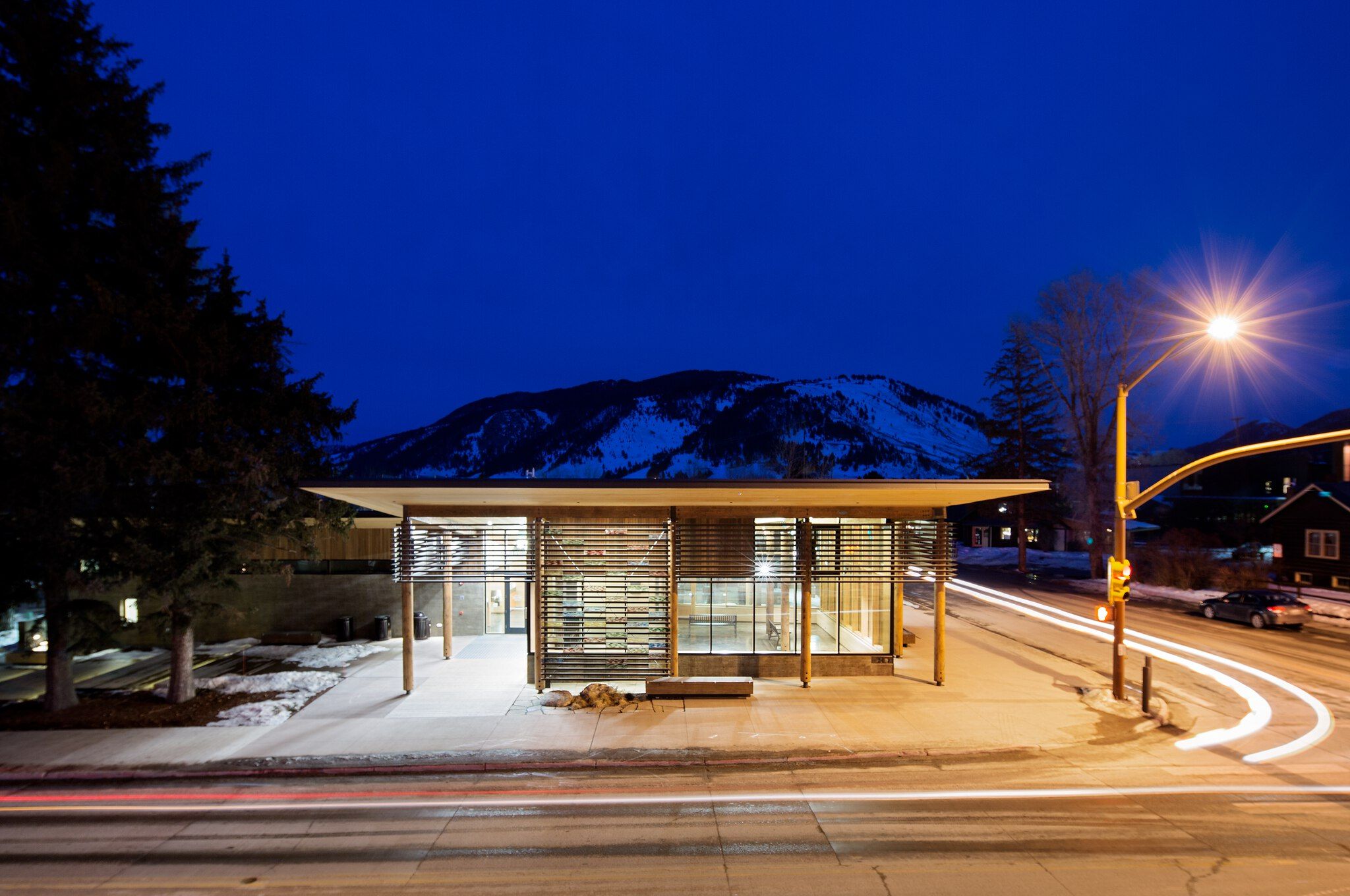 Modern wood and glass building brightly lit at night in a snowy mountain town. Red and white light trails from cars streak across the street in the foreground, with snow-covered mountains silhouetted against a deep blue sky.