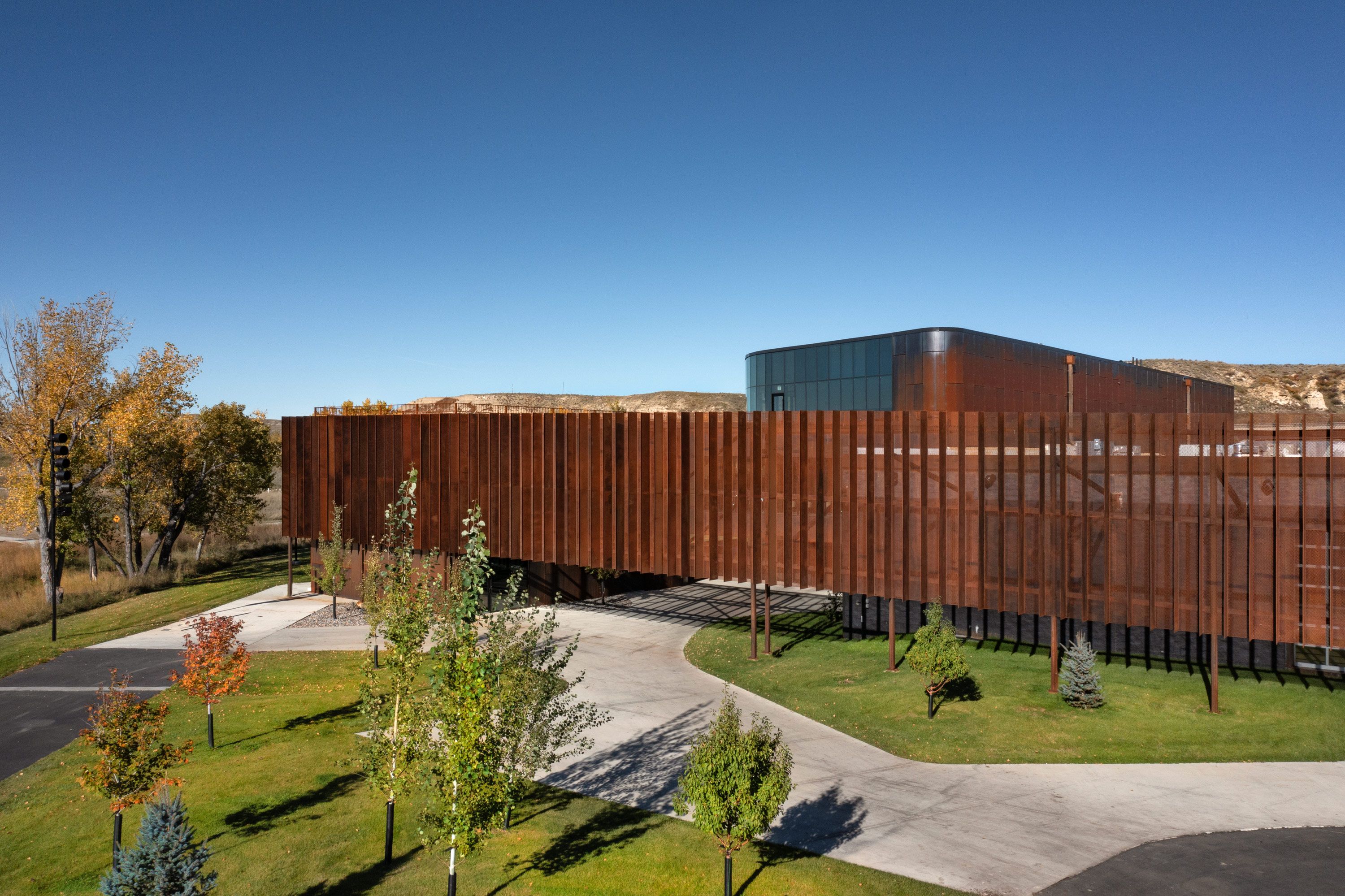 A striking modern building features a facade of rusty brown vertical panels, with a curved glass section visible at one end, all beneath a clear blue sky. The elevated structure casts shadows over green lawns, autumn trees, and a winding paved path in the foreground, while hills frame the distant background.