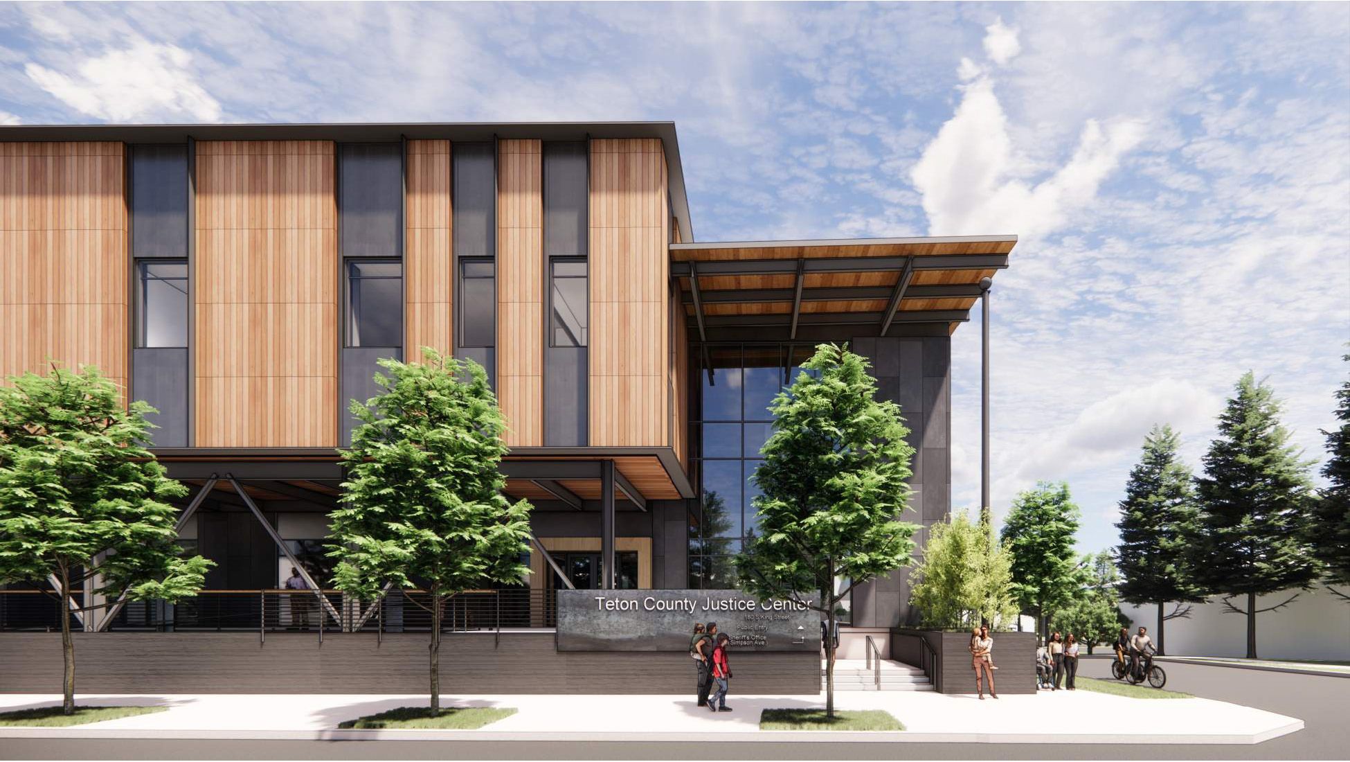 A close-up view of the building's corner and main entrance. A large sign reads "Teton County Justice Center" near a set of concrete stairs and a ramp. The architecture highlights a mix of materials: vertical wood cladding, dark metal supports, and large glass windows. Lush green trees are planted in the foreground and along the street, with a blue sky and light clouds above.