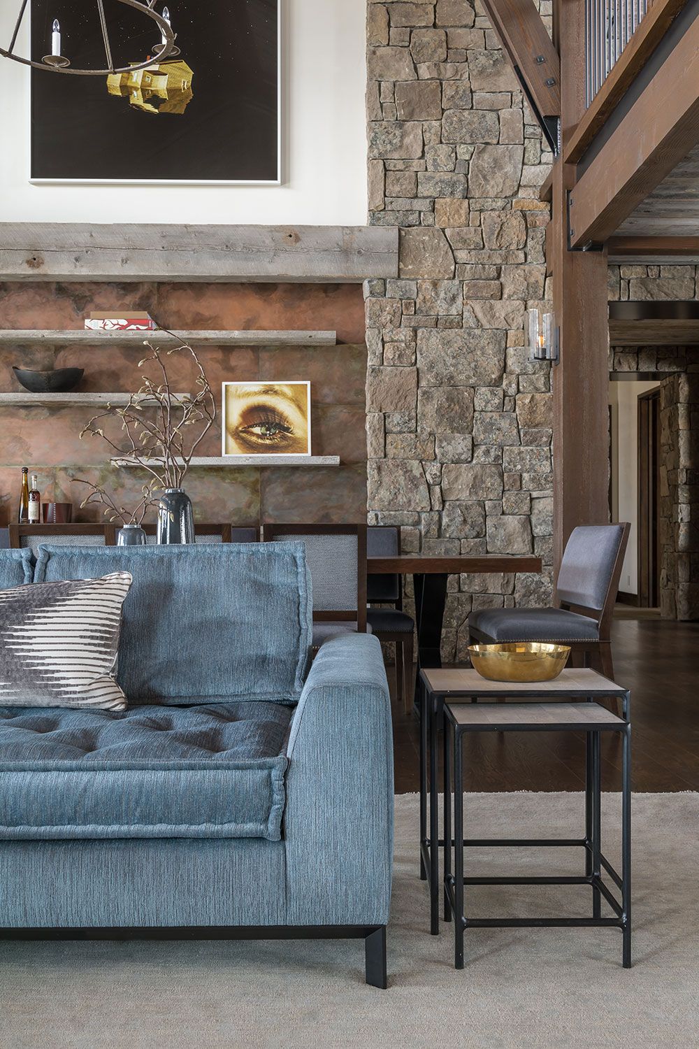 A modern rustic living room featuring a textured blue sofa, stone fireplace pillar, wooden beams, a small nesting side table with a gold bowl, and contemporary artwork on the wall.