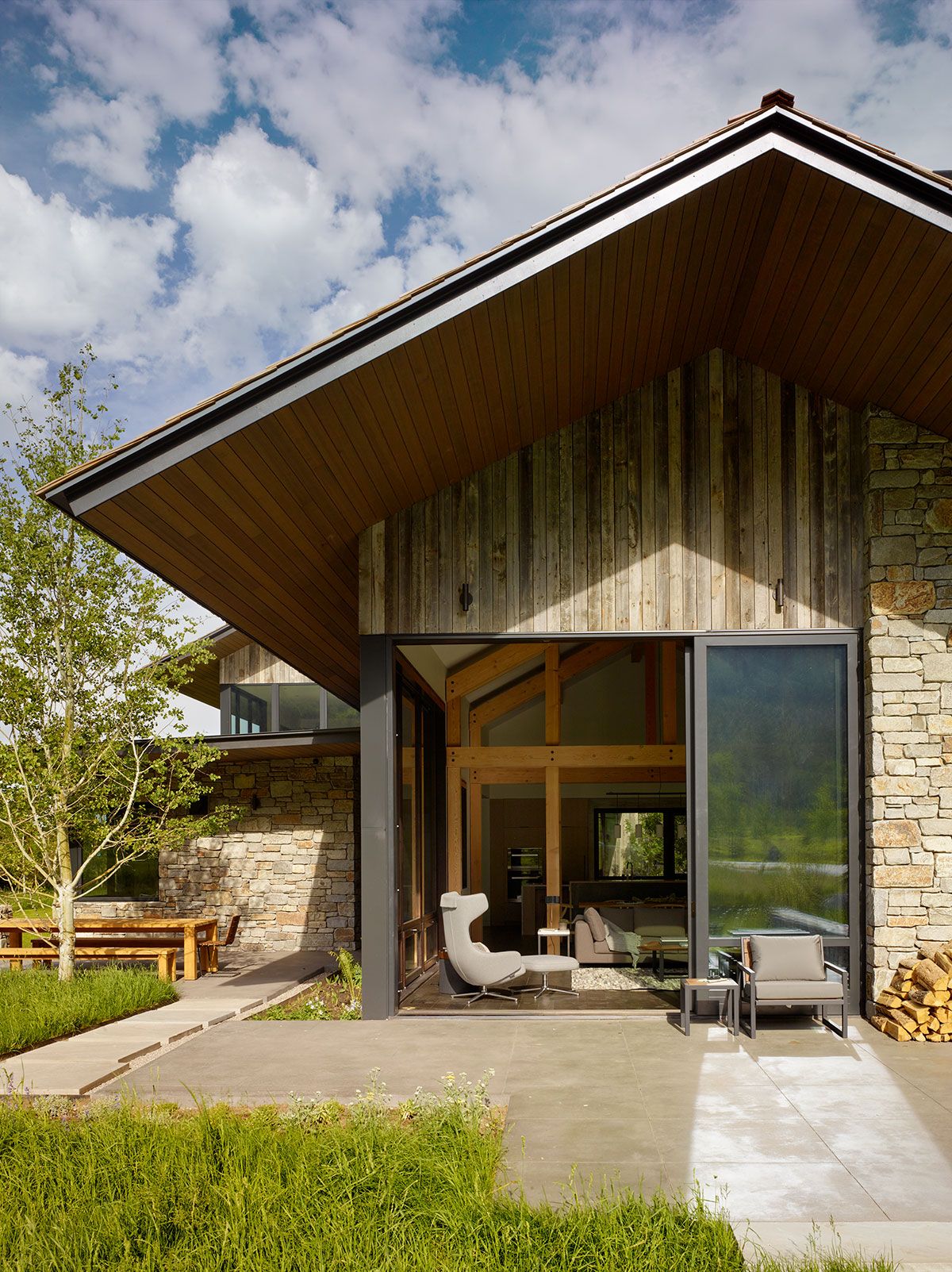 Exterior close-up of a modern rustic house showing a large overhanging wooden roof, natural stone walls, and a wide-open sliding glass door revealing the living area inside.