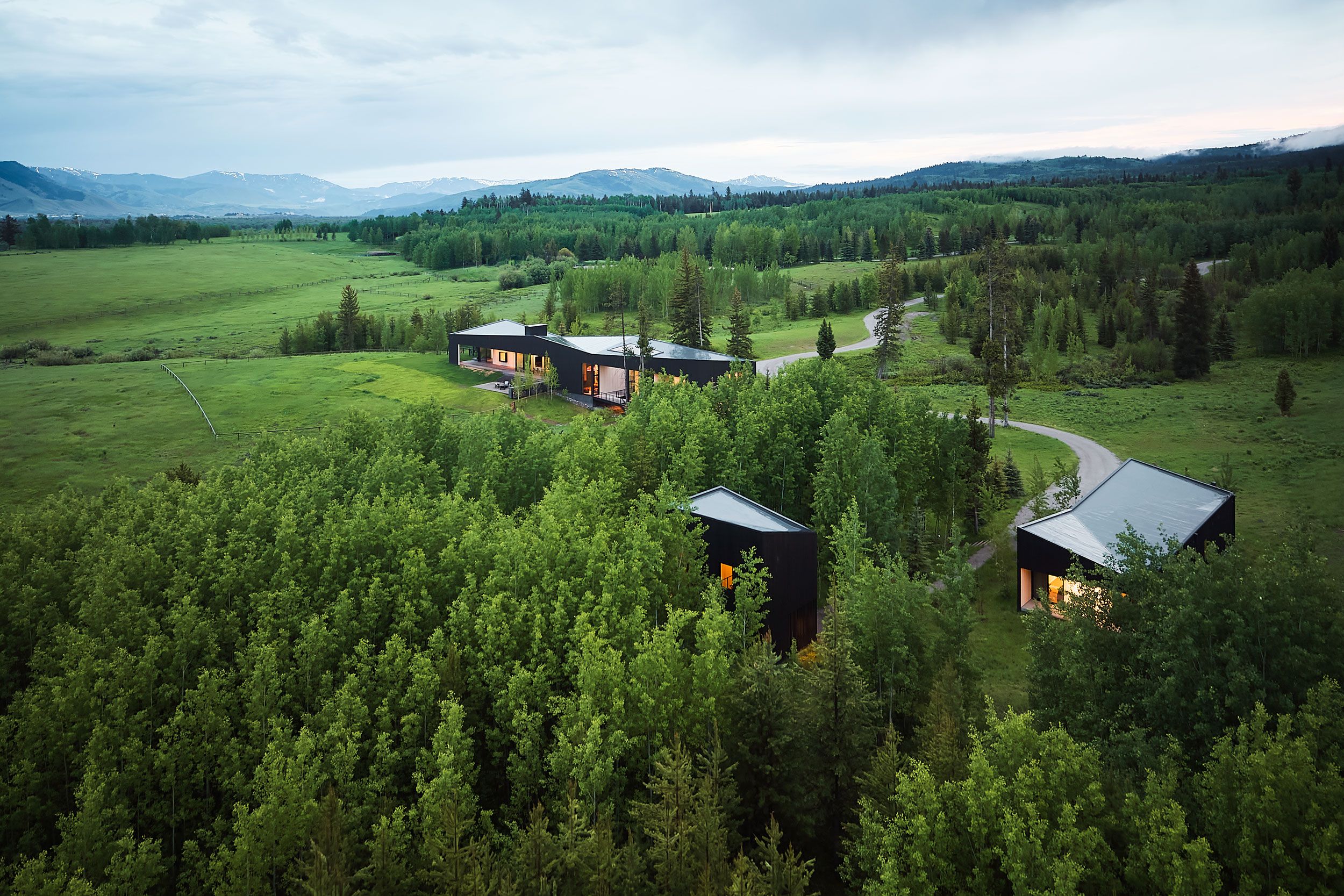 Aerial view of a modern black architectural complex with geometric roofs, nestled in a lush green forest. Interior lights glow from the buildings, with winding paths leading to open fields and distant snow-capped mountains under an overcast sky.