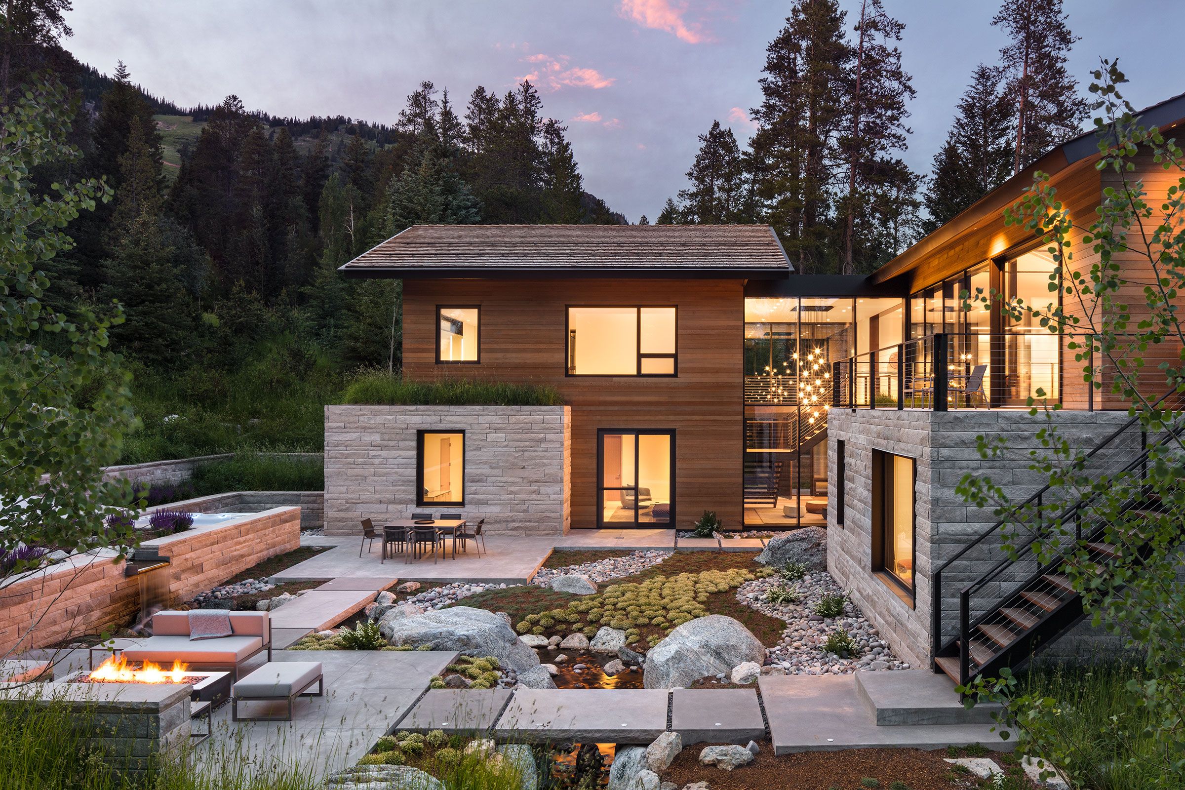 Evening view of a luxurious modern home's illuminated backyard courtyard, showcasing a stone-wrapped patio, fire pit lounge area, a small stream, and glowing interior lights.