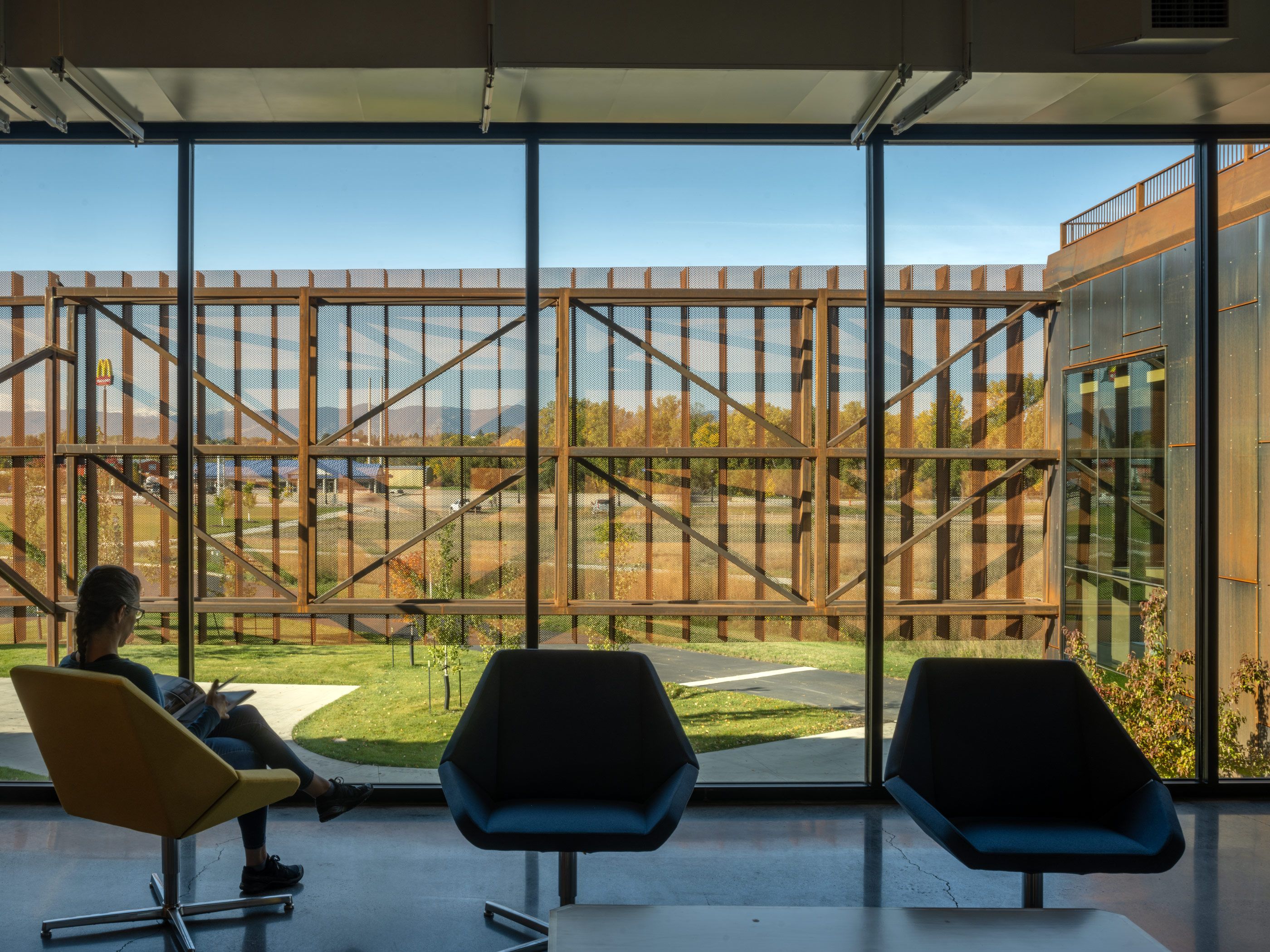 Woman reading in a yellow lounge chair by a large window overlooking an architectural screen of weathered metal. The outdoor view includes autumn trees, distant mountains, and a McDonald's sign under a clear blue sky. Two empty dark chairs sit nearby.