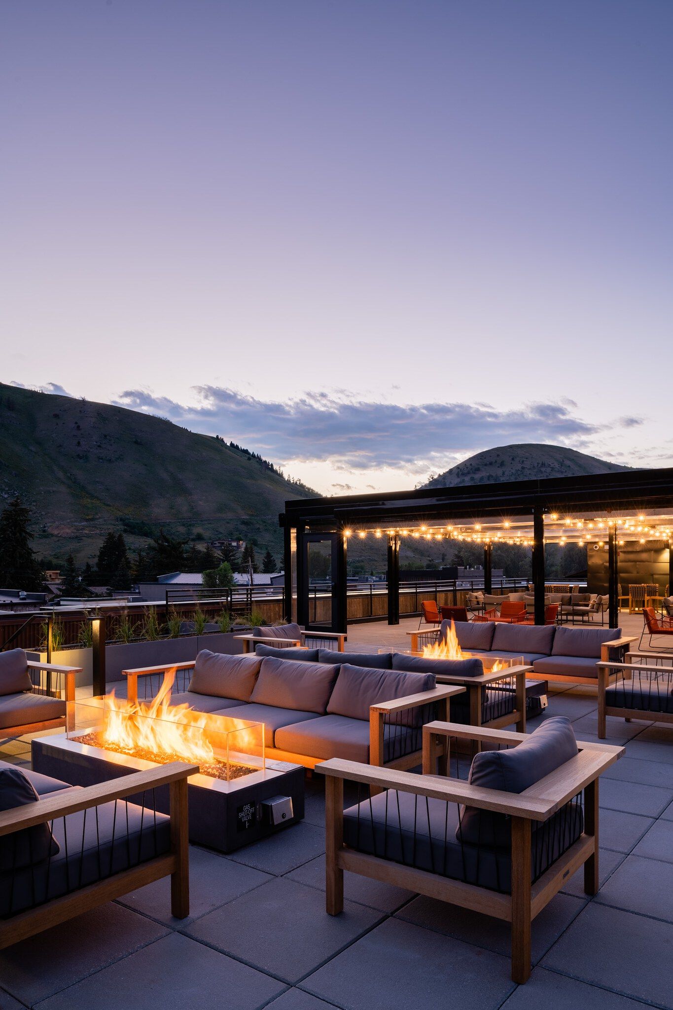Inviting rooftop lounge at dusk with multiple fire pits, modern outdoor sofas, and string lights, overlooking a mountain landscape.